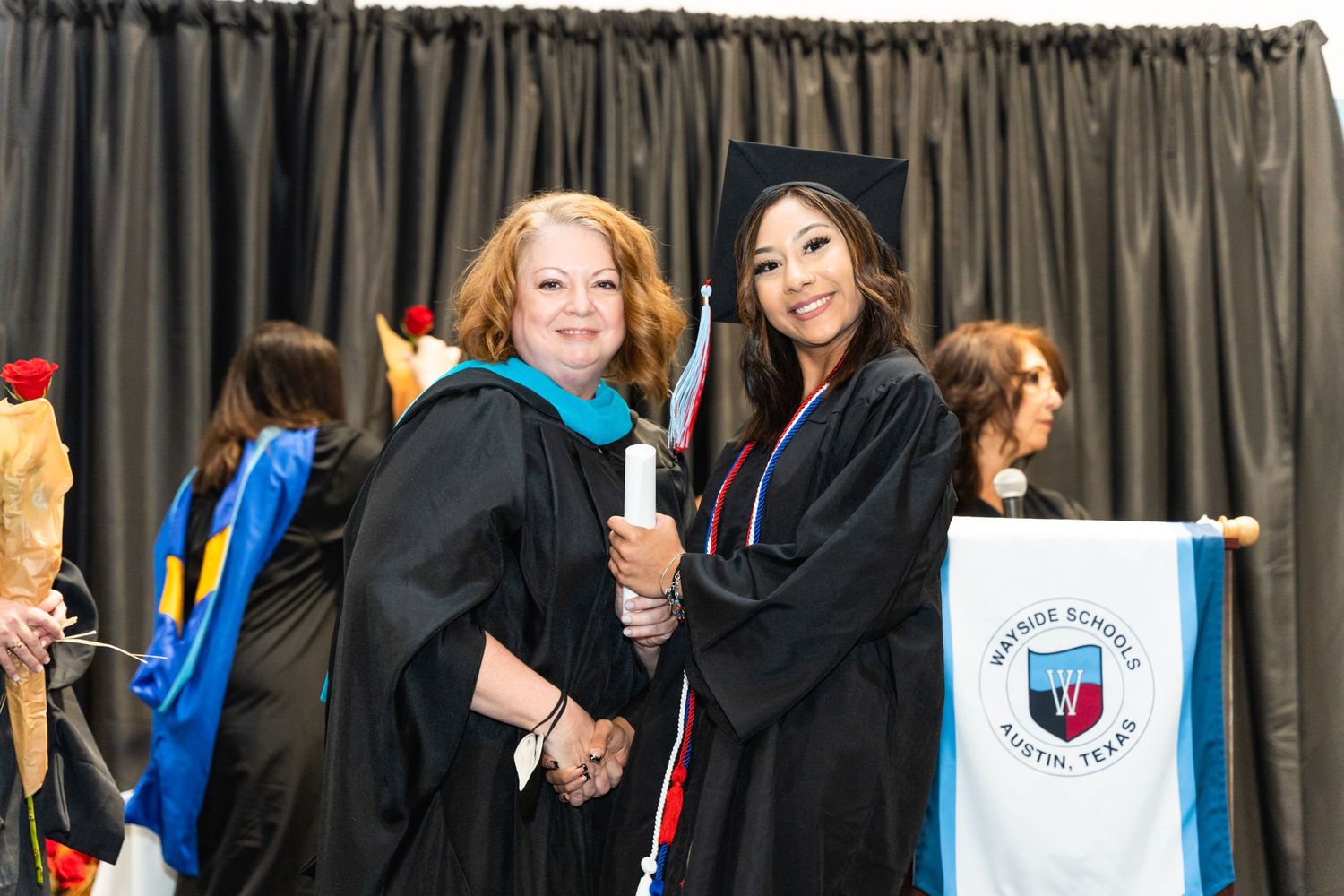 Two graduates in black caps and gowns pose together at commencement ceremony.