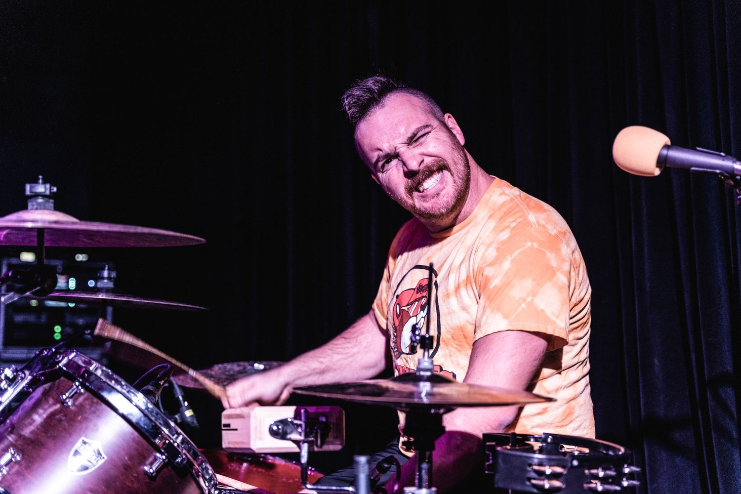 Close up of drummer playing and smiling during performance under orange stage lighting.