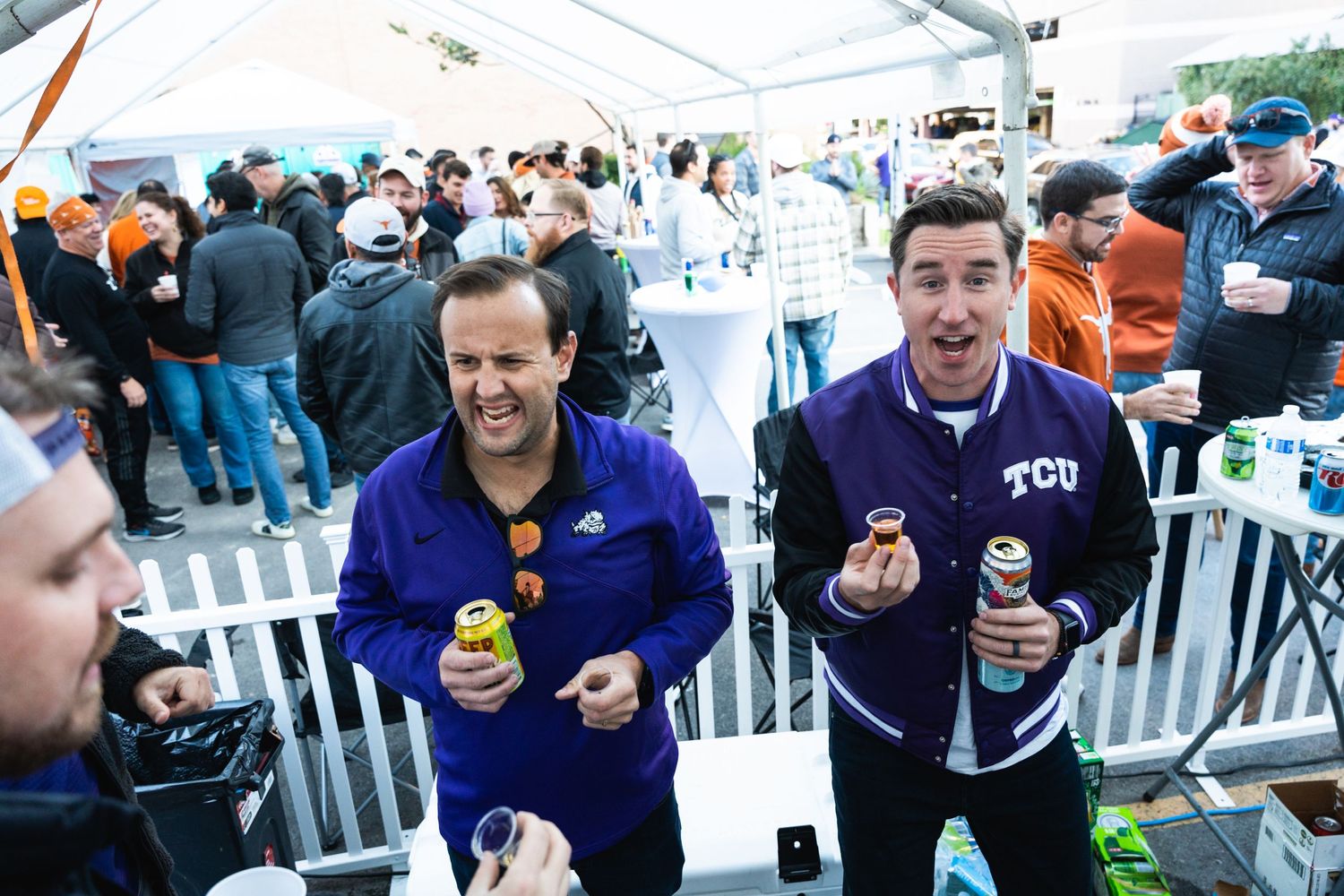 Two fans in purple jerseys enjoying beverages at an outdoor sports event.