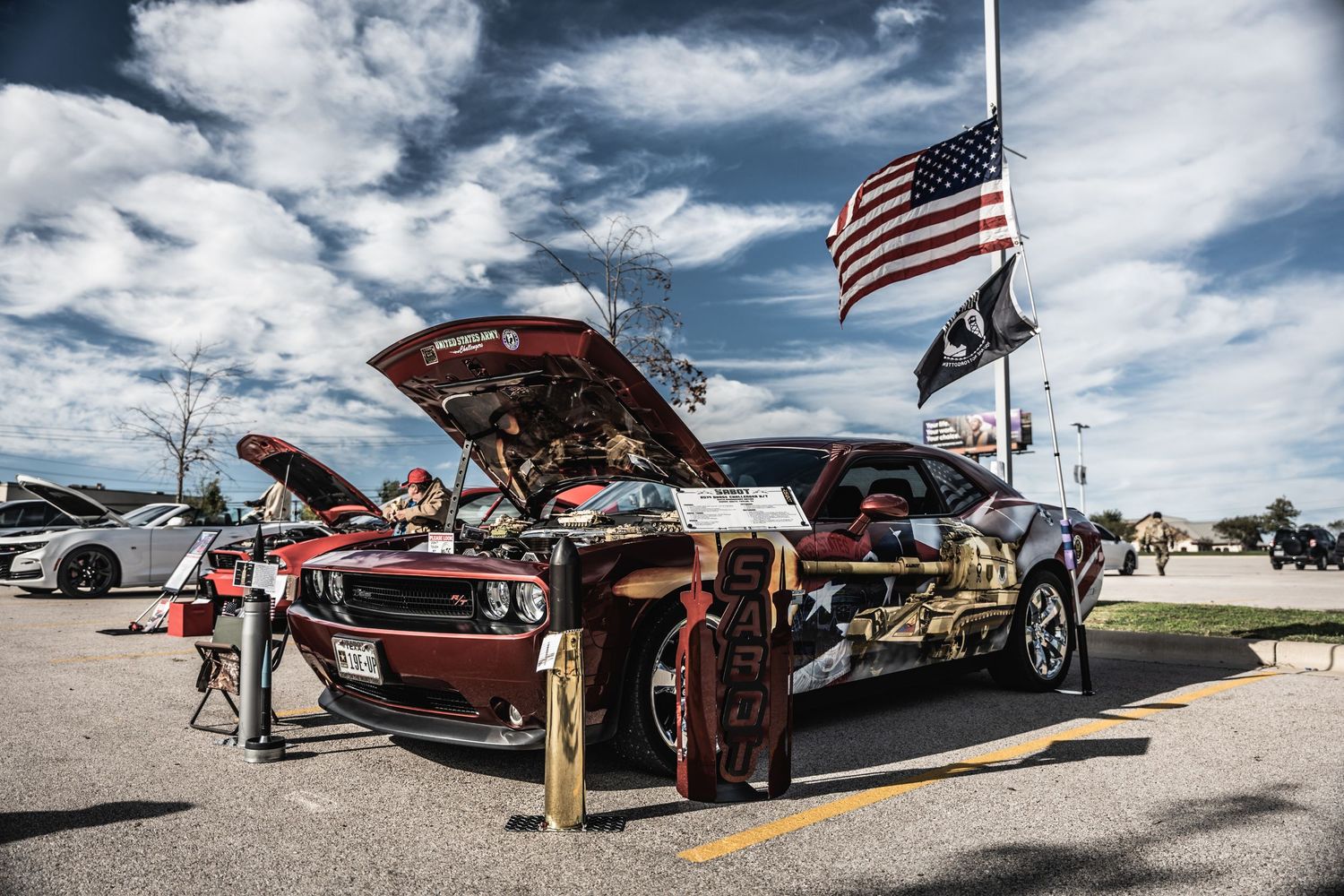 Classic muscle cars on display at outdoor car show with American flags flying overhead.
