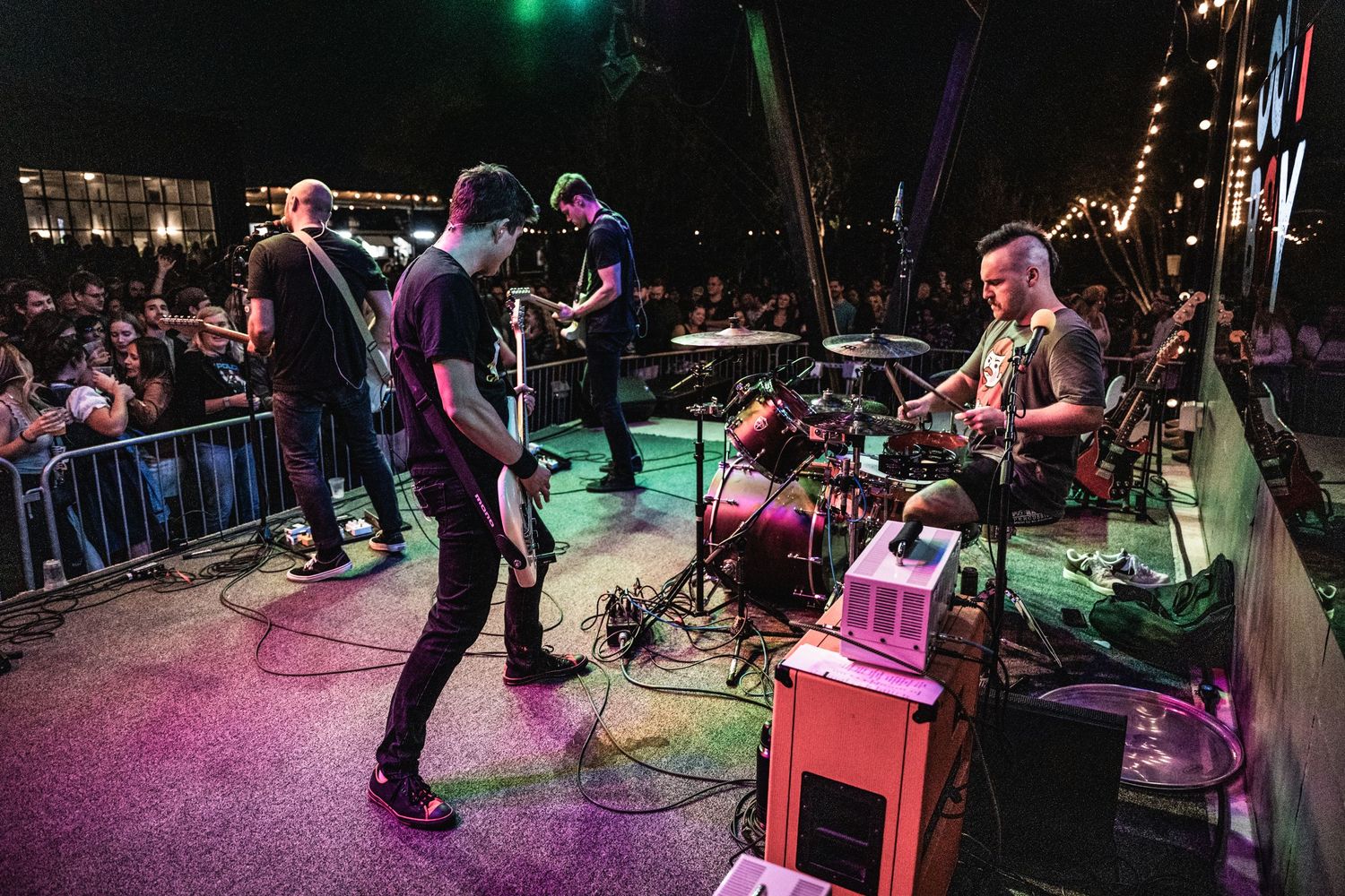 Band performing under colorful stage lights with crowd visible behind barricades.