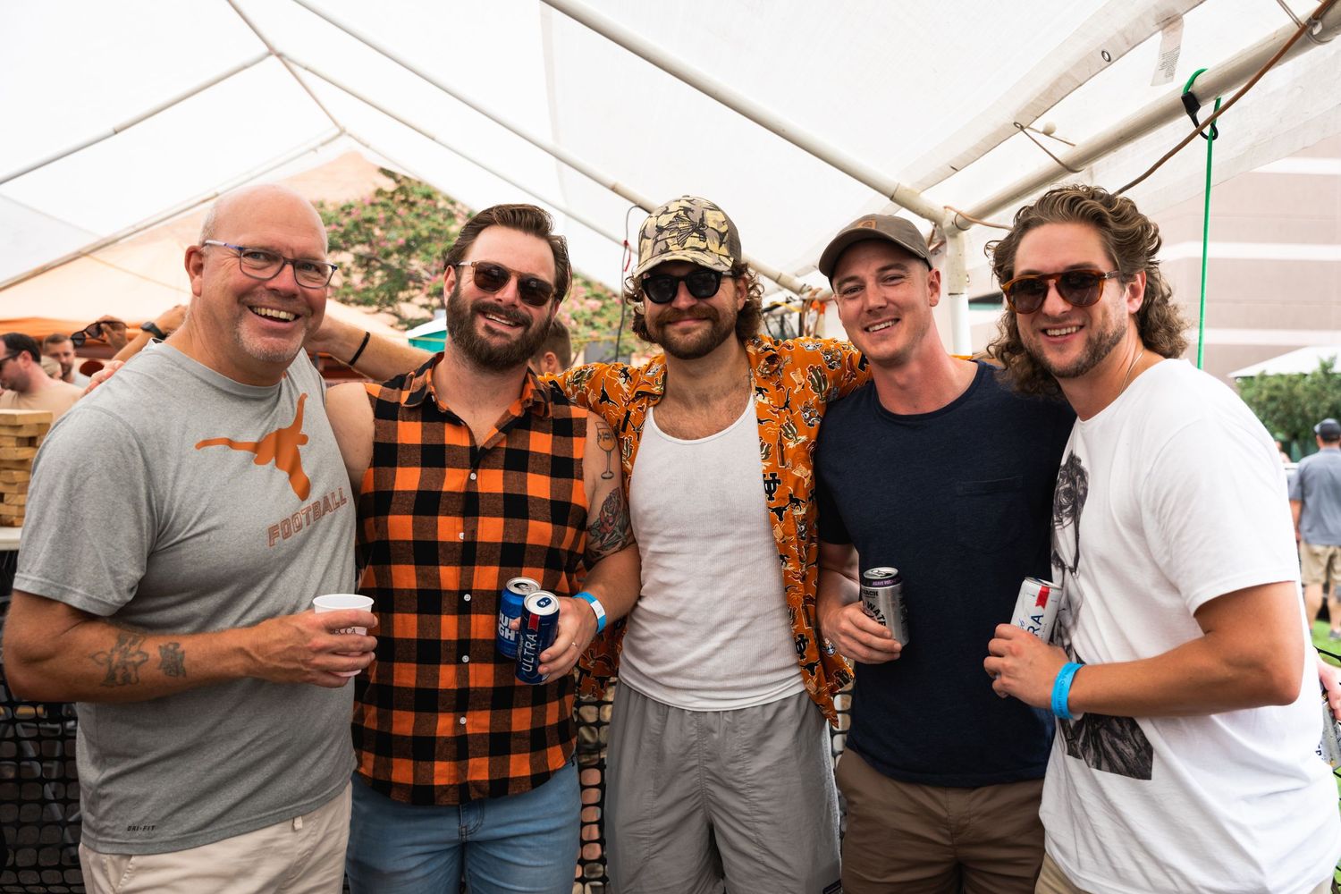 Five friends posing together with drinks at an outdoor festival.