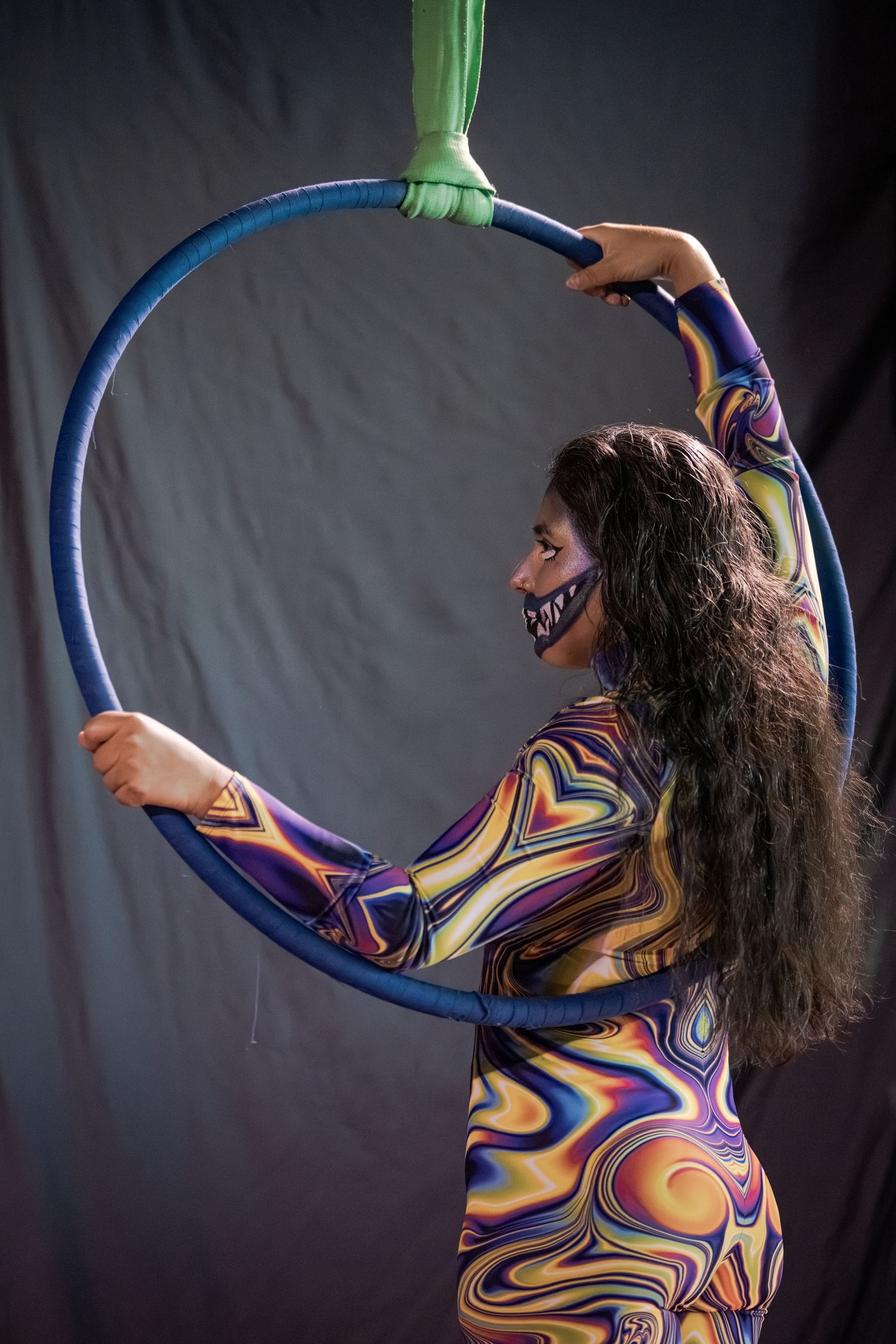 A female aerial hoop performer in a colorful psychedelic bodysuit and decorative mask poses gracefully with a blue lyra ring.