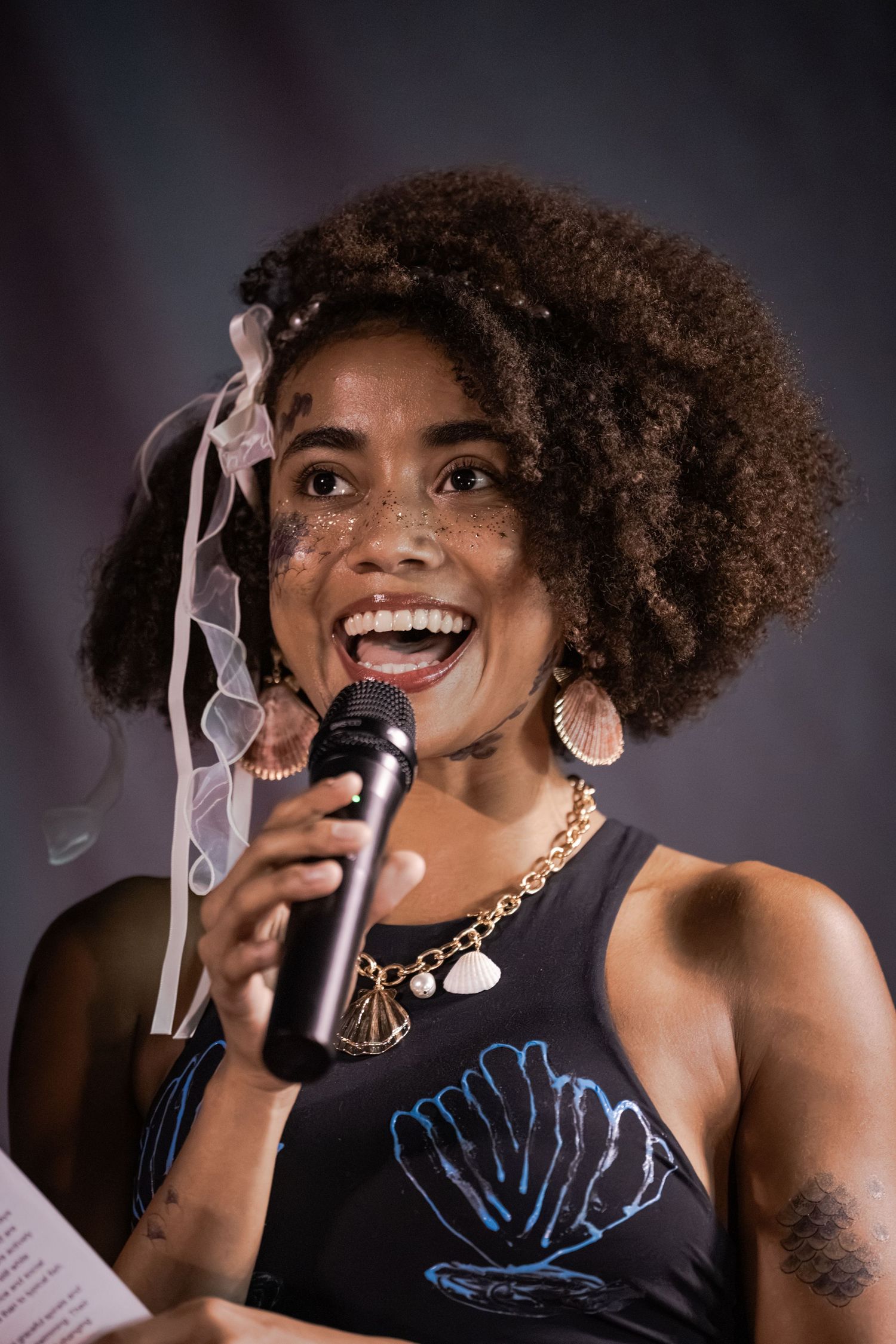 Joyful woman with curly hair and freckles singing into a microphone, wearing a black top and gold necklace.