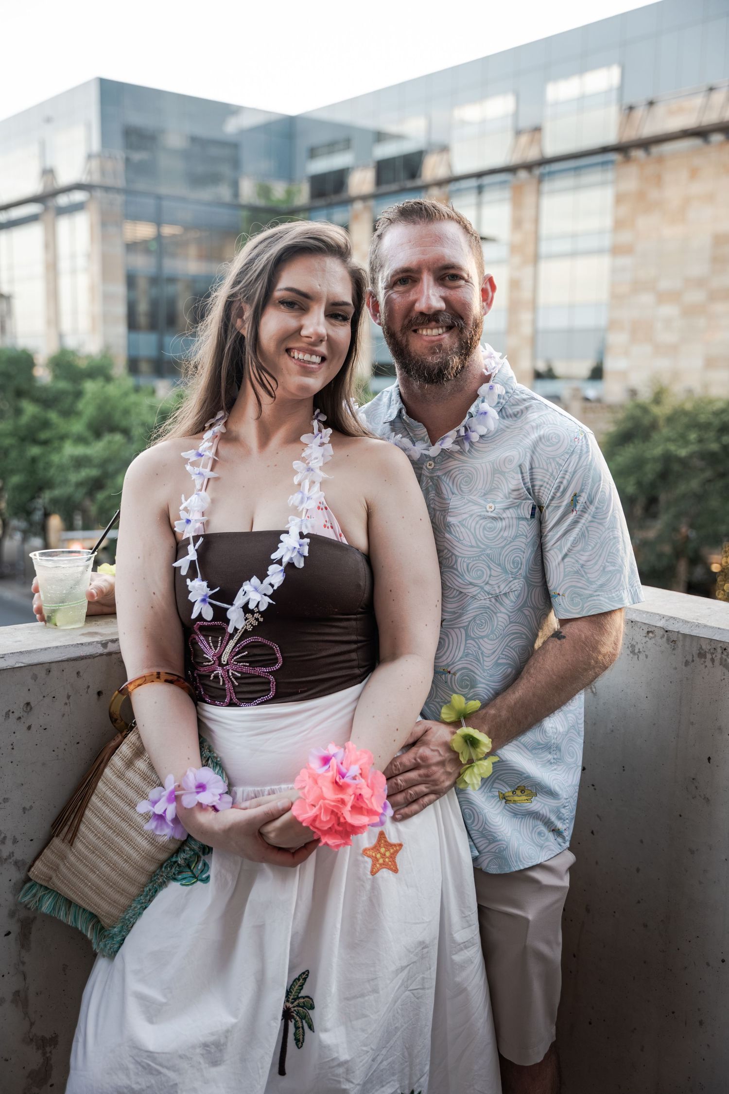 Couple wearing floral leis smiling at a tropical-themed outdoor party with a modern building backdrop.