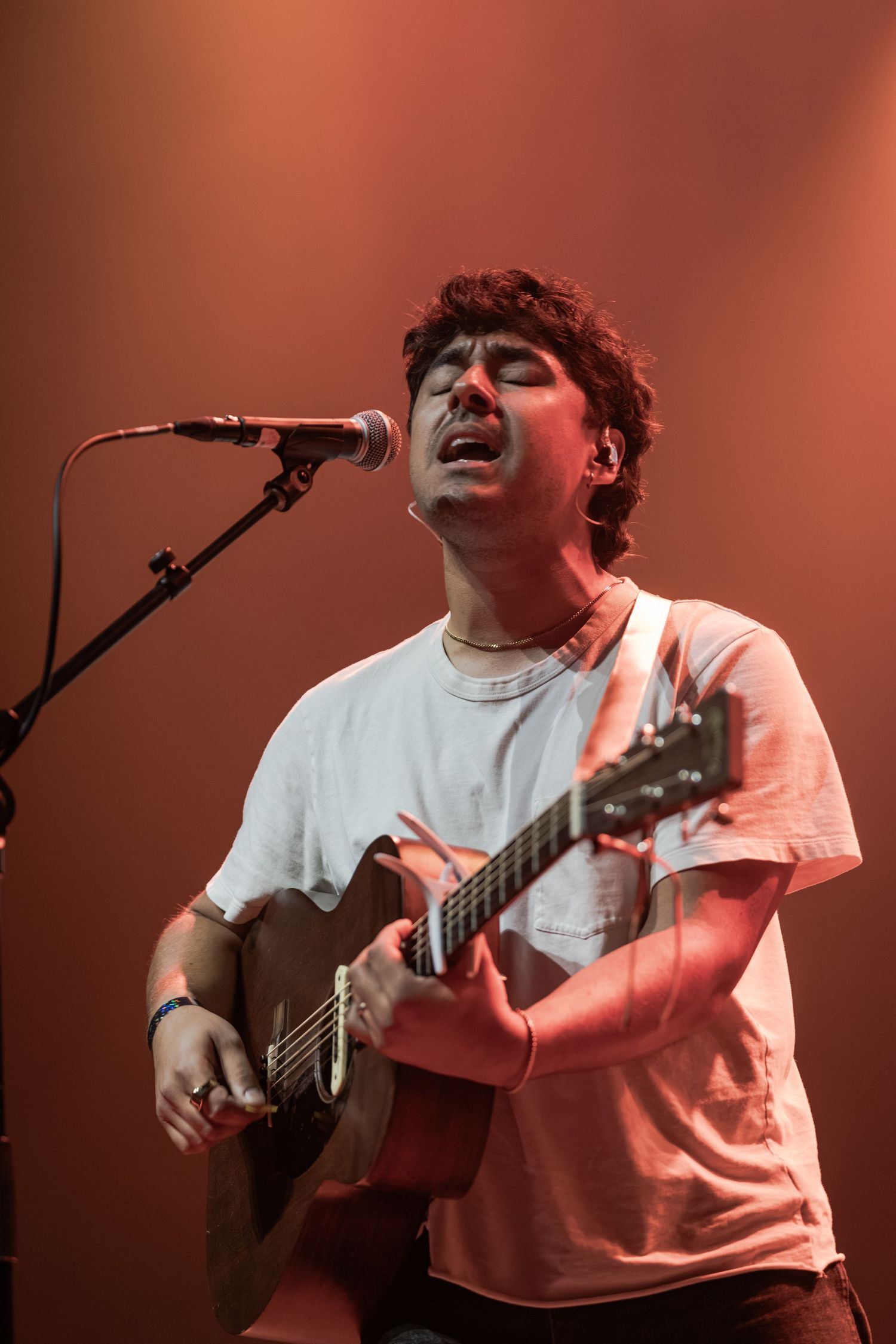 Young male musician plays acoustic guitar and sings passionately into microphone on stage under warm red lighting.