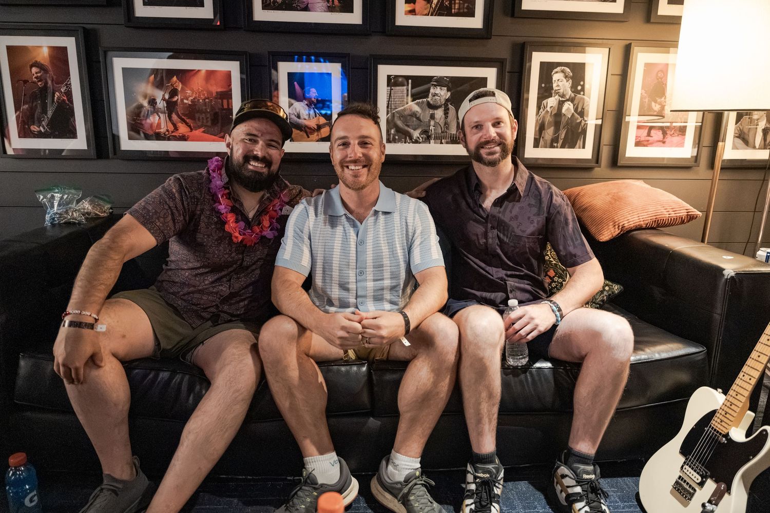 Three smiling men sitting on a black couch in a music venue surrounded by framed concert photos.