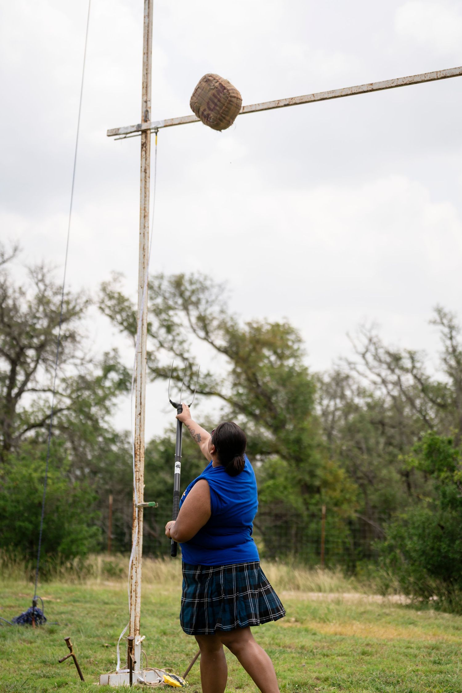 A person in a blue top and plaid skirt attempts to hit a target on a tall wooden pole in a grassy field.