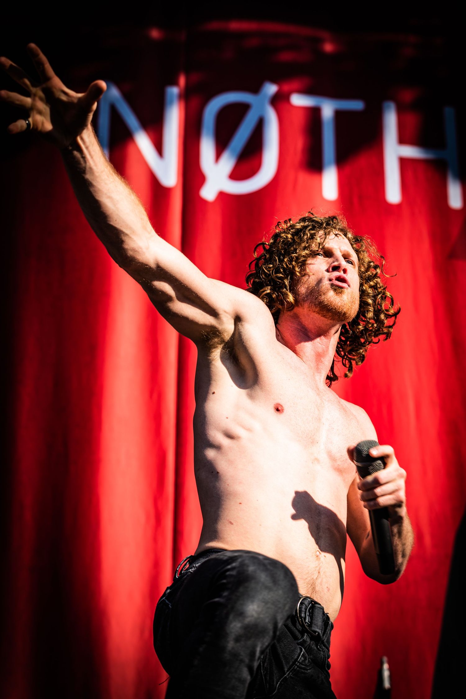 Shirtless performer sings passionately on stage with red curtain backdrop in dramatic lighting.