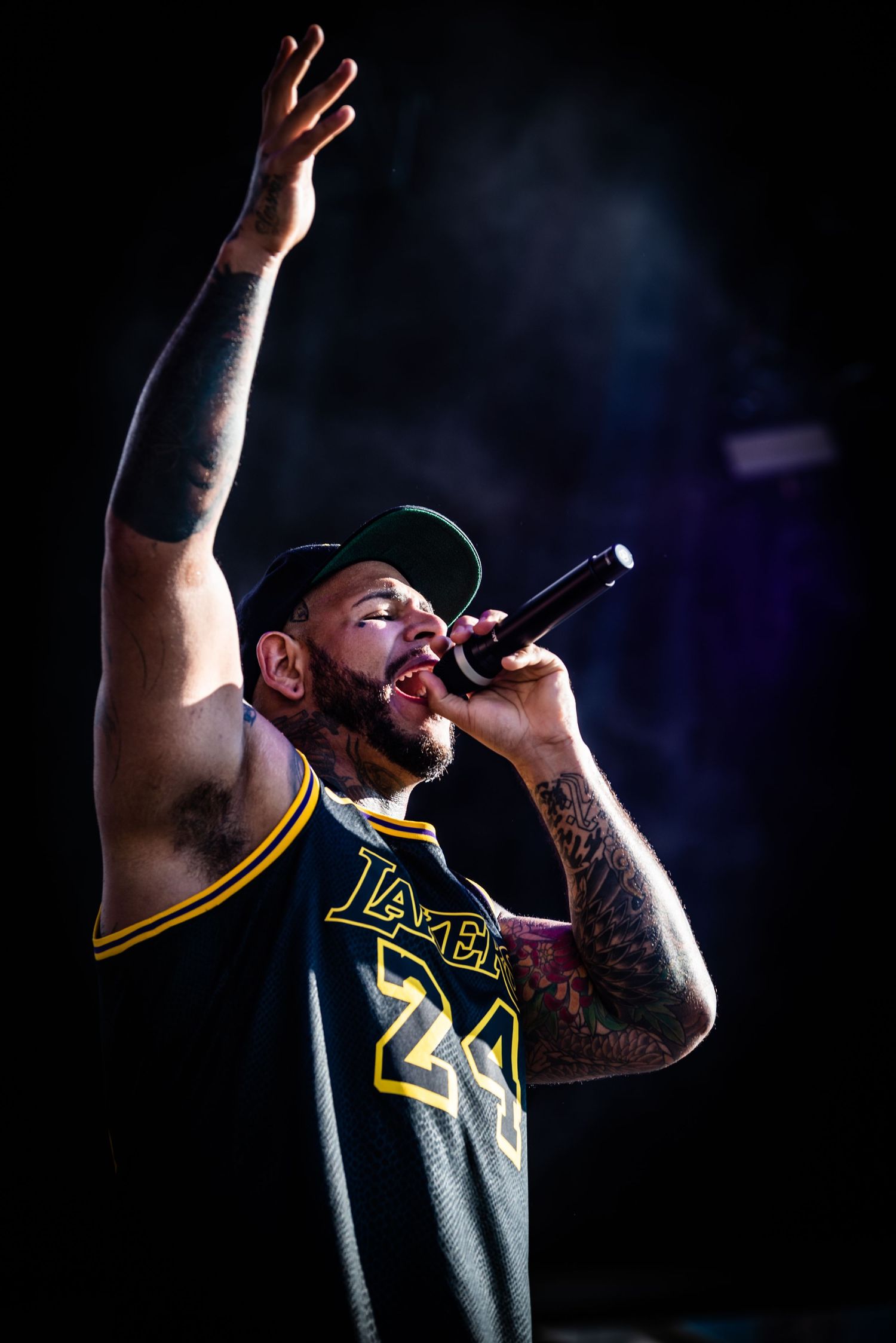 Performer wearing Lakers jersey number 24 holds microphone and raises arm while singing on dark stage.