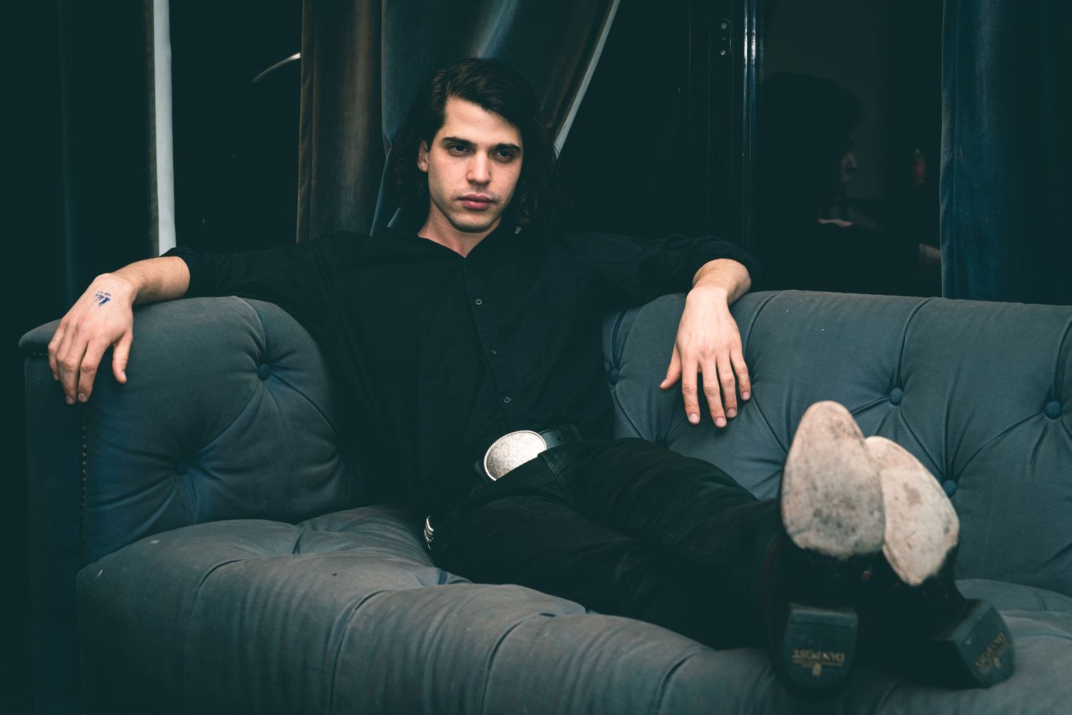 A person wearing black clothing sits casually on a dark tufted couch in a dimly lit room.