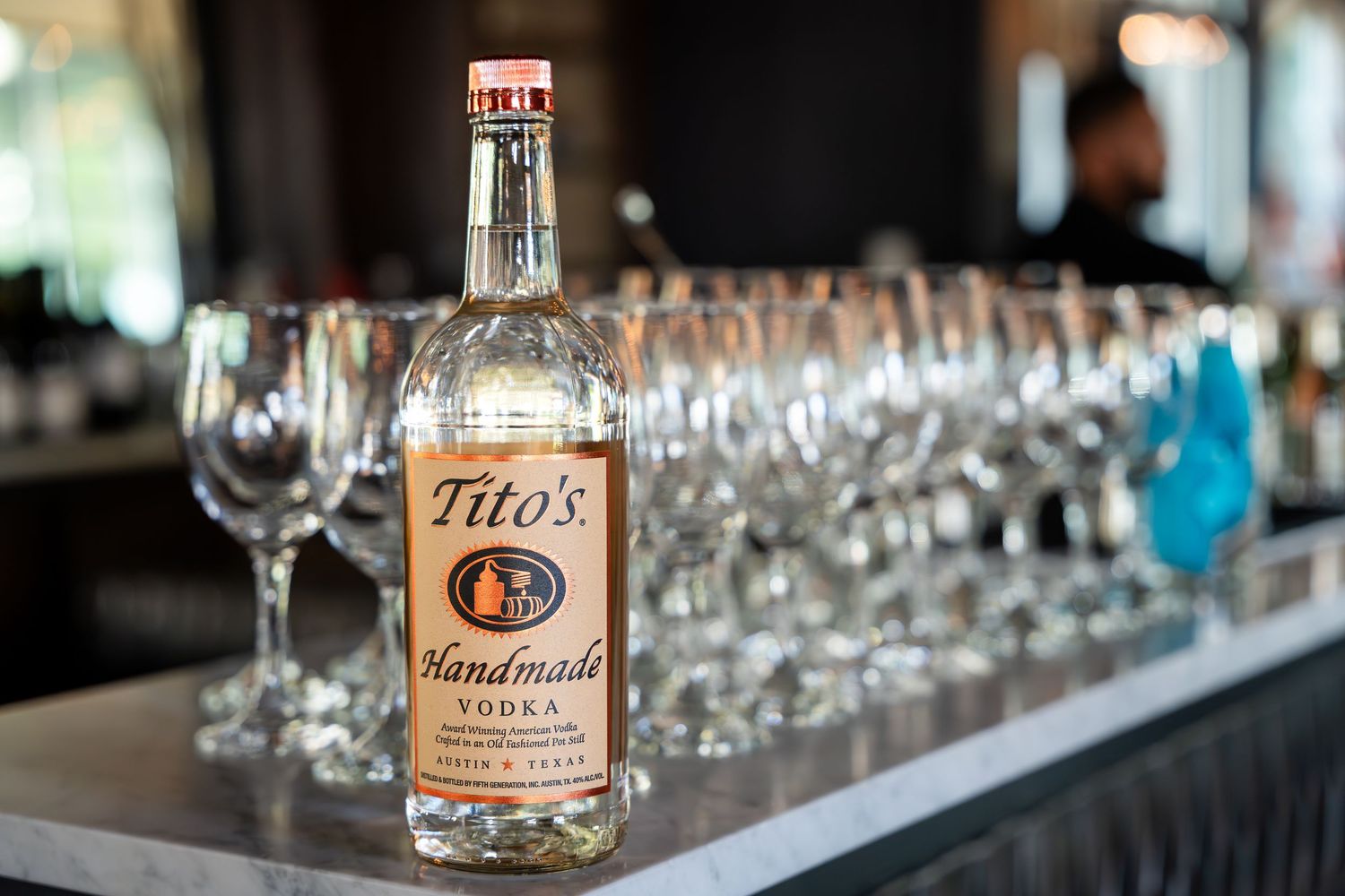A row of glass bottles and wine glasses arranged on a bar counter with soft lighting and blurred background.
