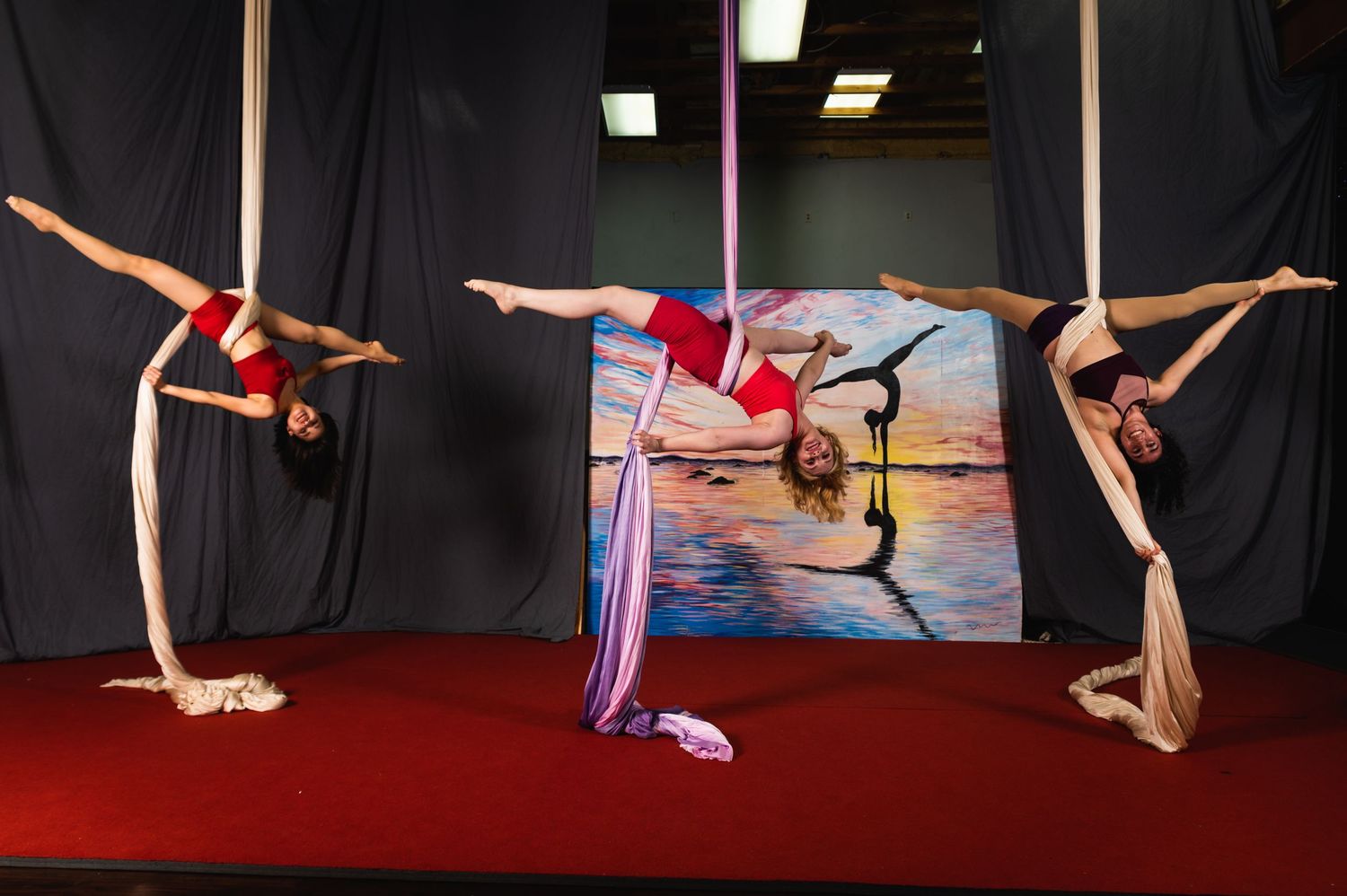 Aerial dancers perform graceful poses while suspended from red fabric in a dark studio space.