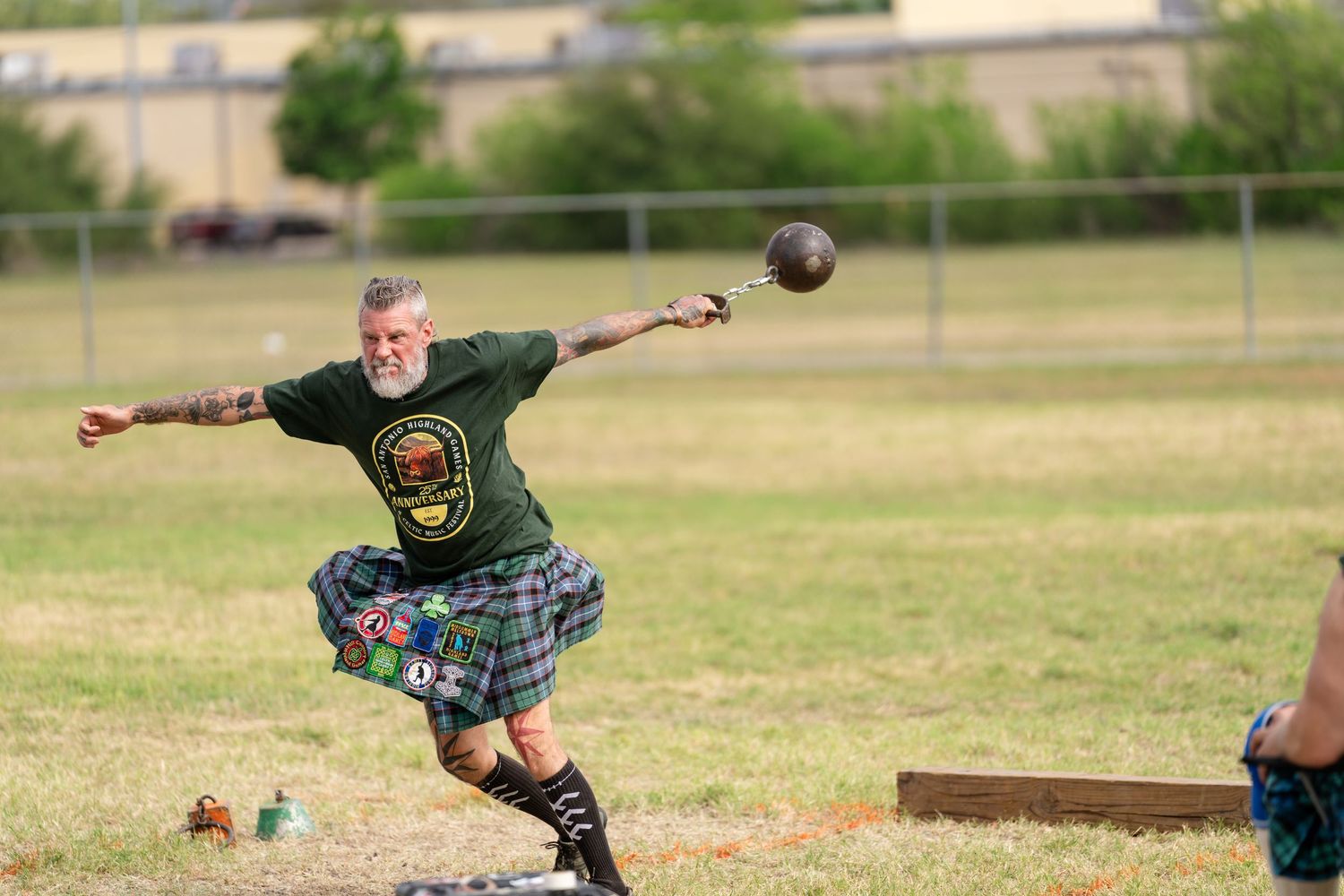 Tattooed athlete in green shirt and tartan kilt throws heavy ball at Highland Games competition outdoors.