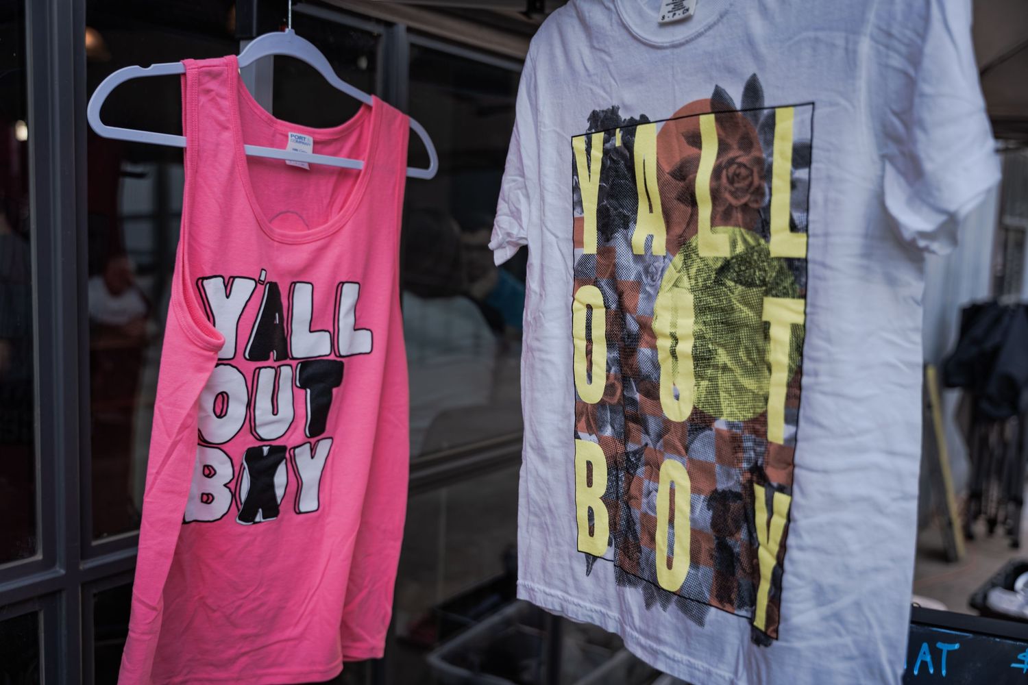 Two Fall Out Boy band t-shirts hanging on display, a pink tank top and white tee with colorful graphic print.