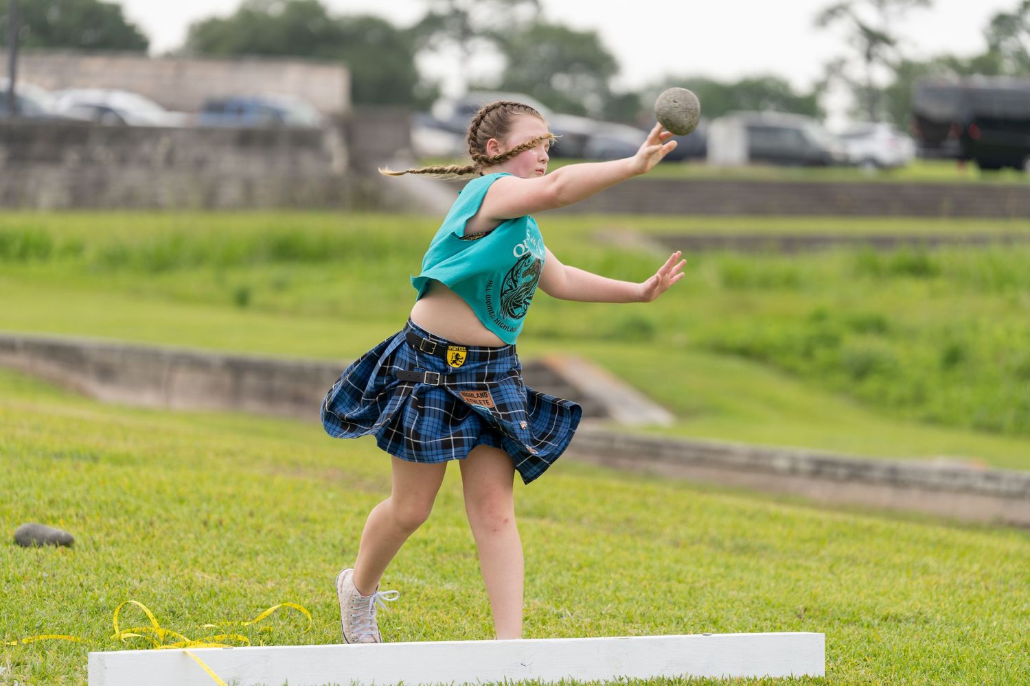 Young girl in tartan skirt throwing shot put at Highland Games competition on green grass field.