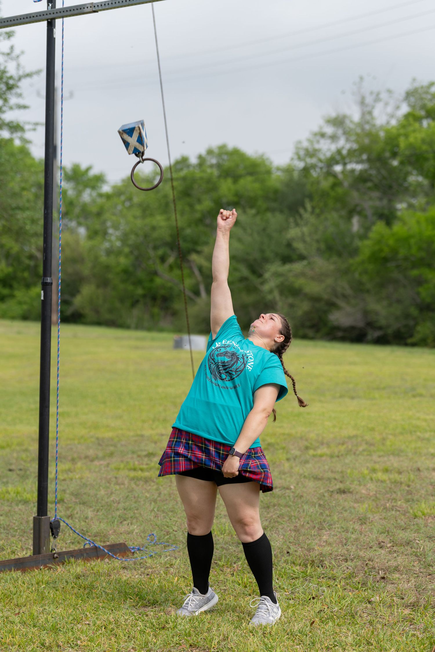 Woman in tartan skirt and teal shirt competing in Scottish Highland Games weight over bar event outdoors.