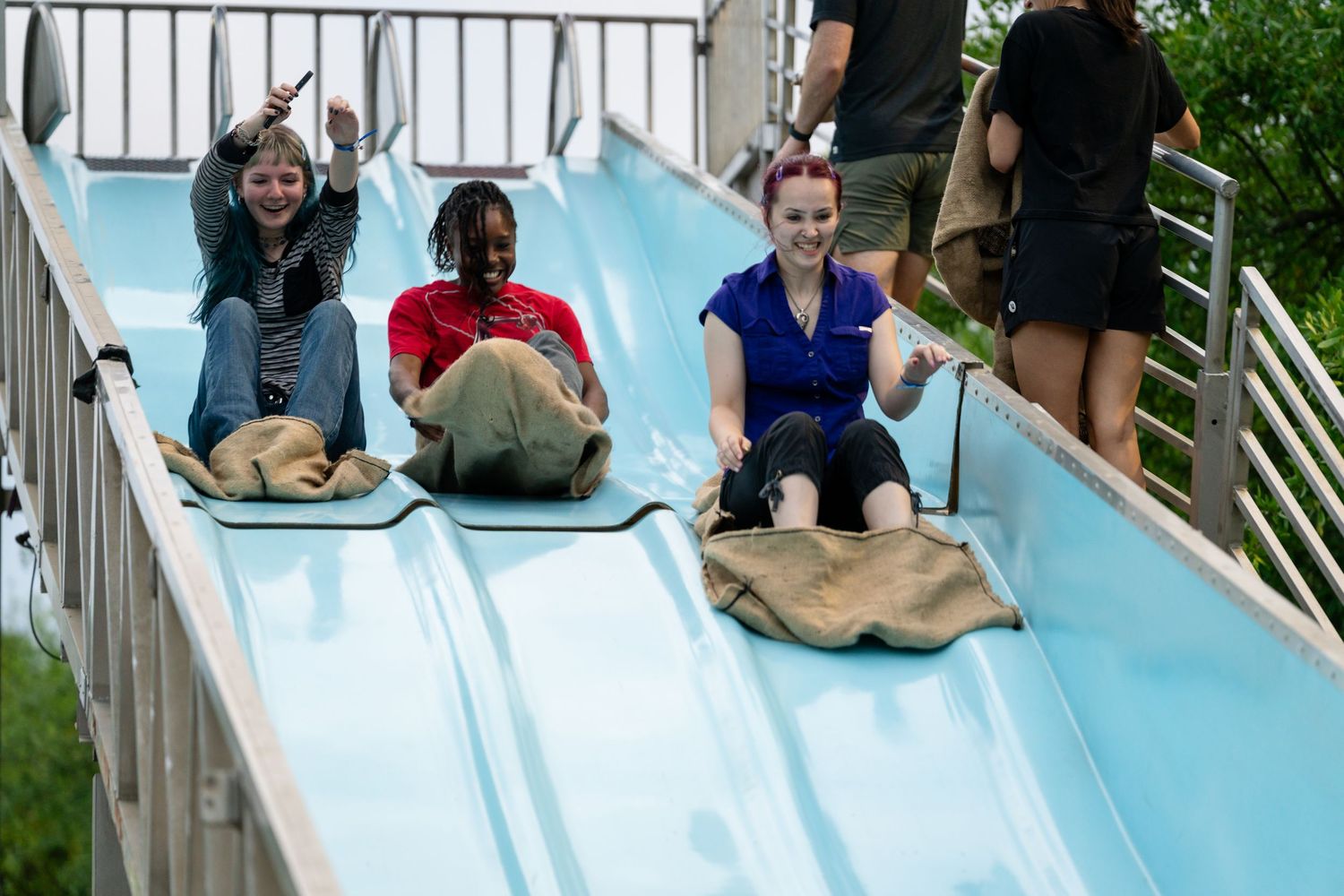 Three people smiling and sliding down a blue amusement park slide on burlap sacks on a sunny day.