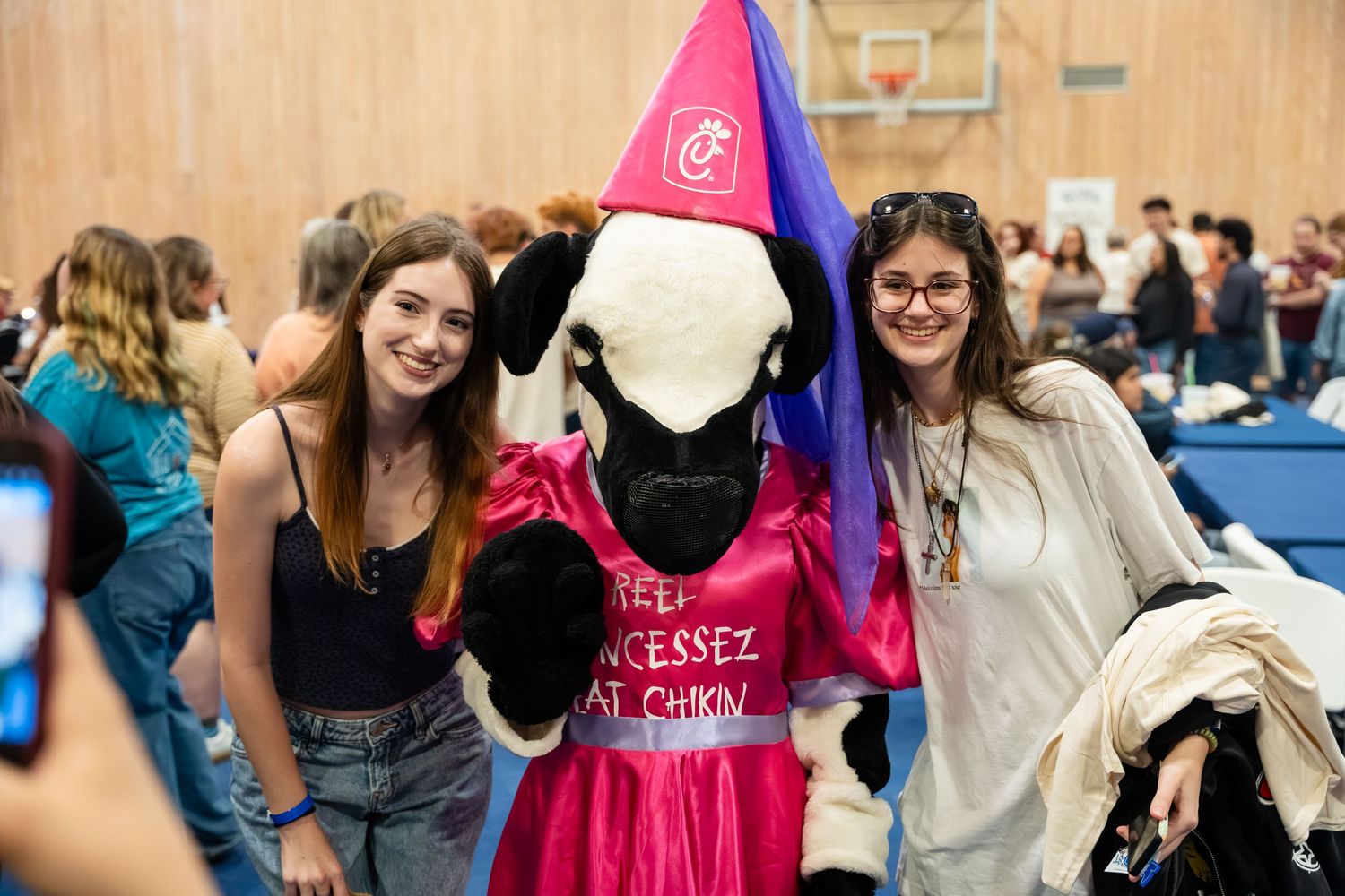 Two students pose with a Chick-fil-A cow mascot in a wizard costume at a college event.