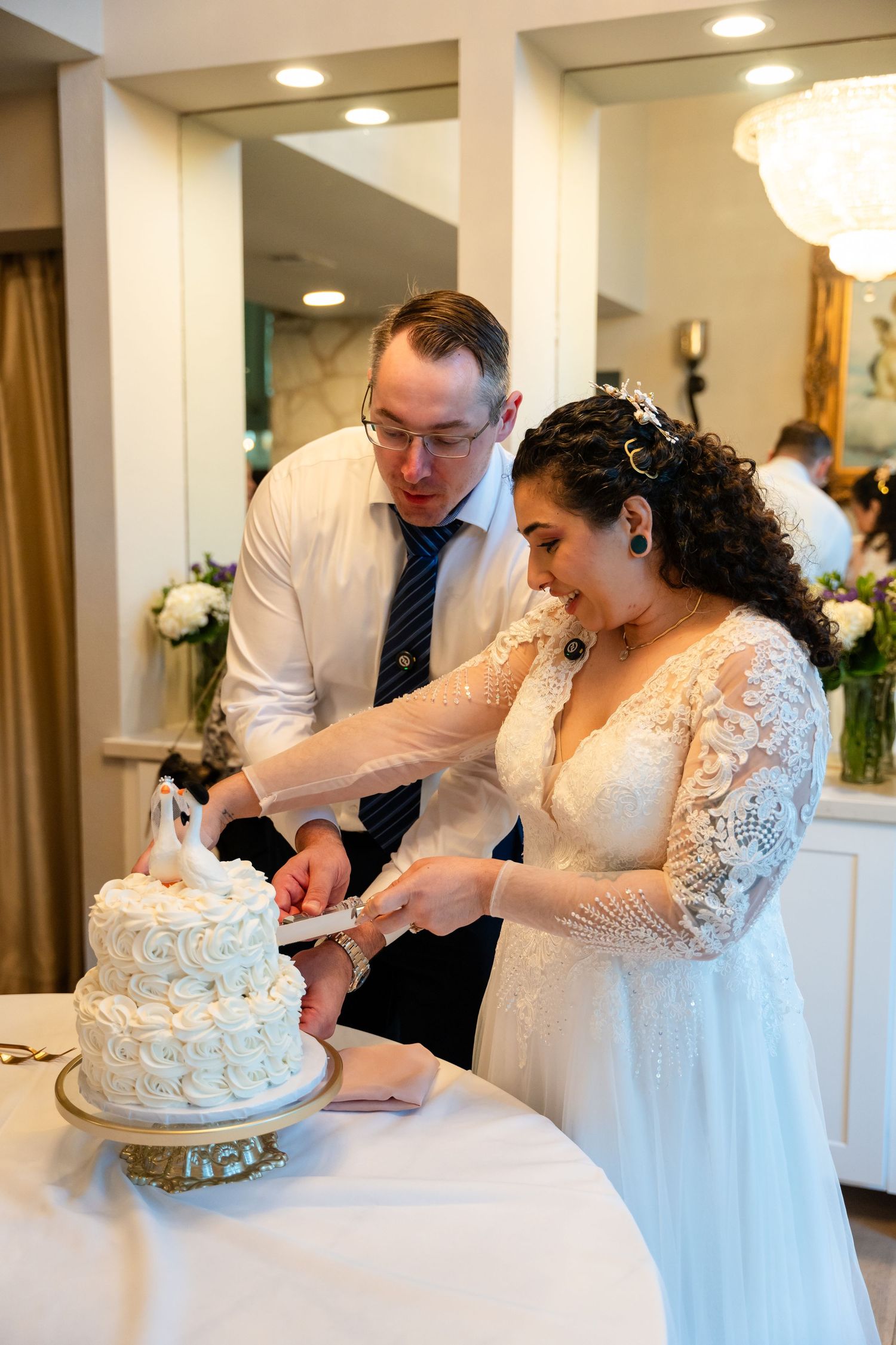 Bride and groom cutting white rosette wedding cake together at indoor wedding reception celebration.