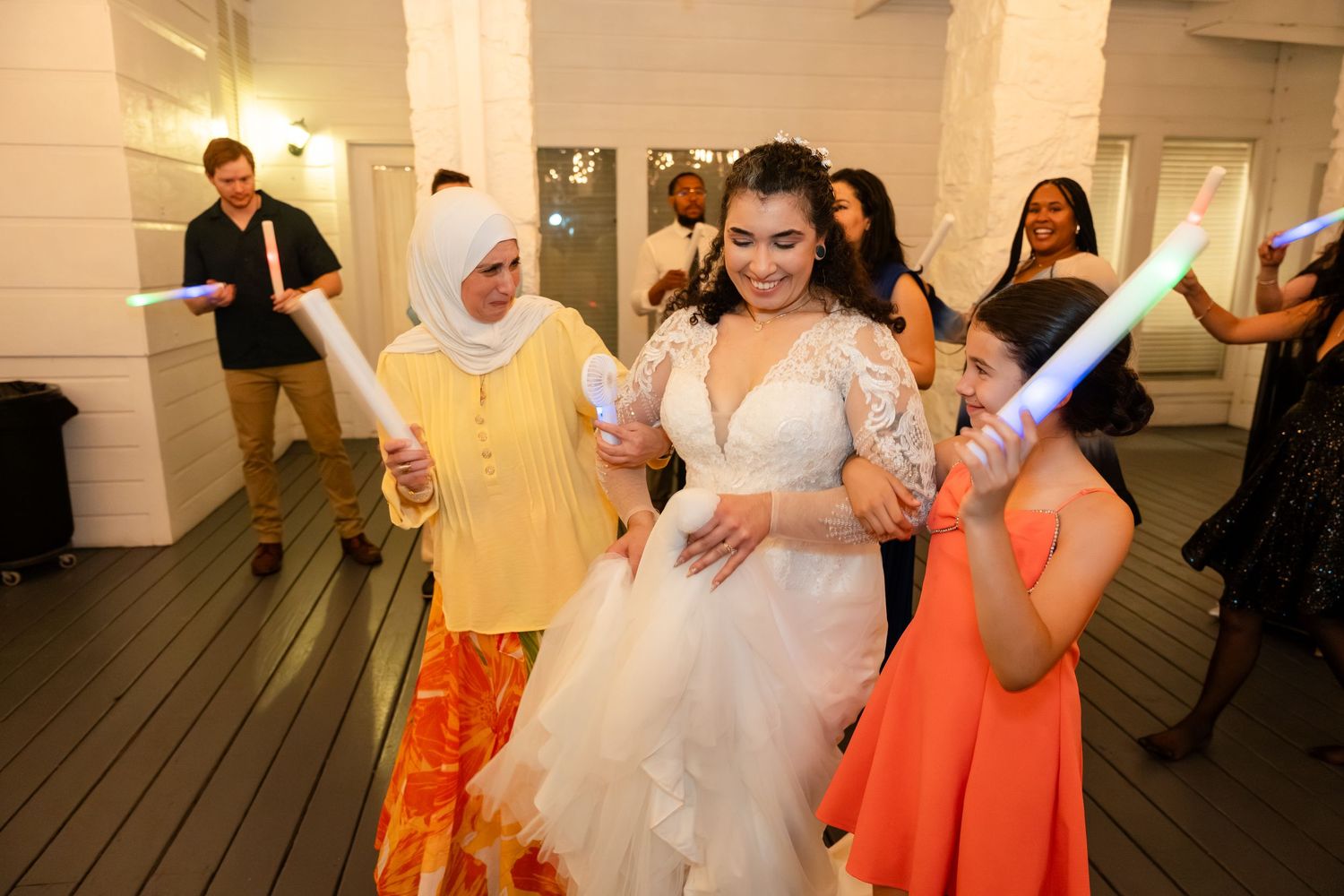 Bride in white wedding gown dancing with guests holding colorful glow sticks at a festive wedding reception.