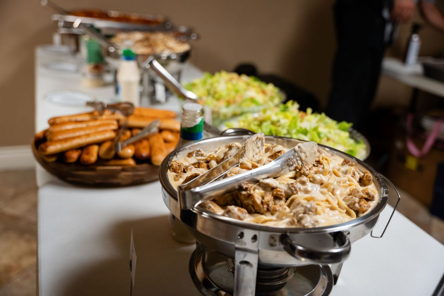 Buffet table with chafing dish of creamy chicken, spring rolls, and fresh green salad at a catered event.