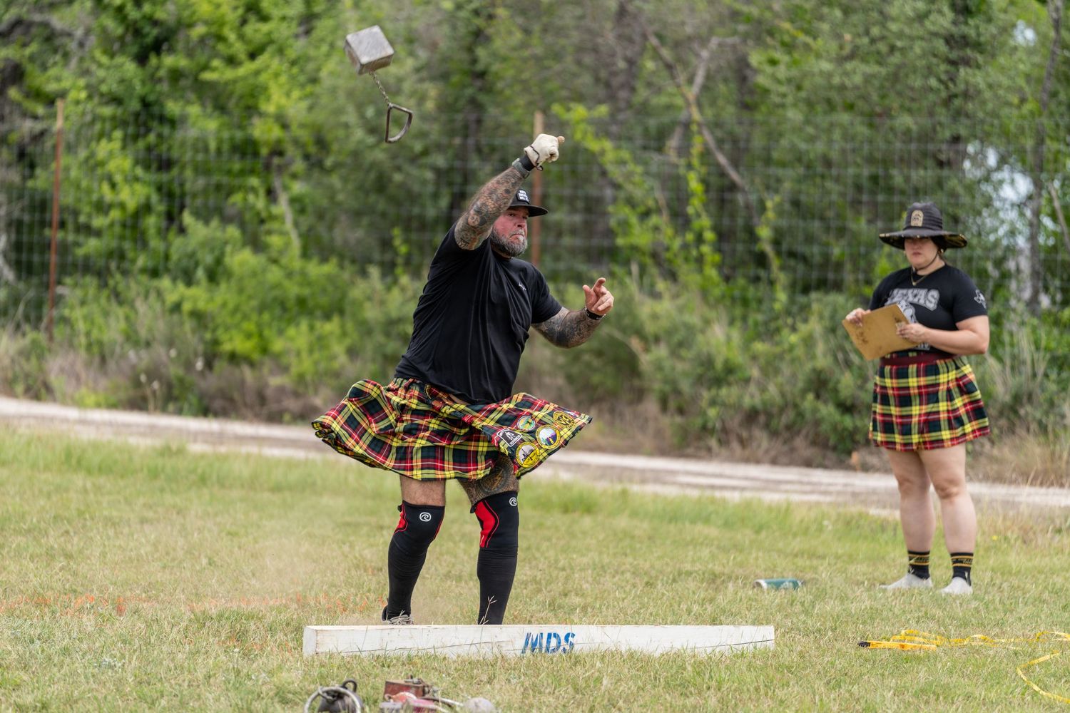 Two men in colorful kilts compete in Highland Games hammer throw on a sunny day outdoors.