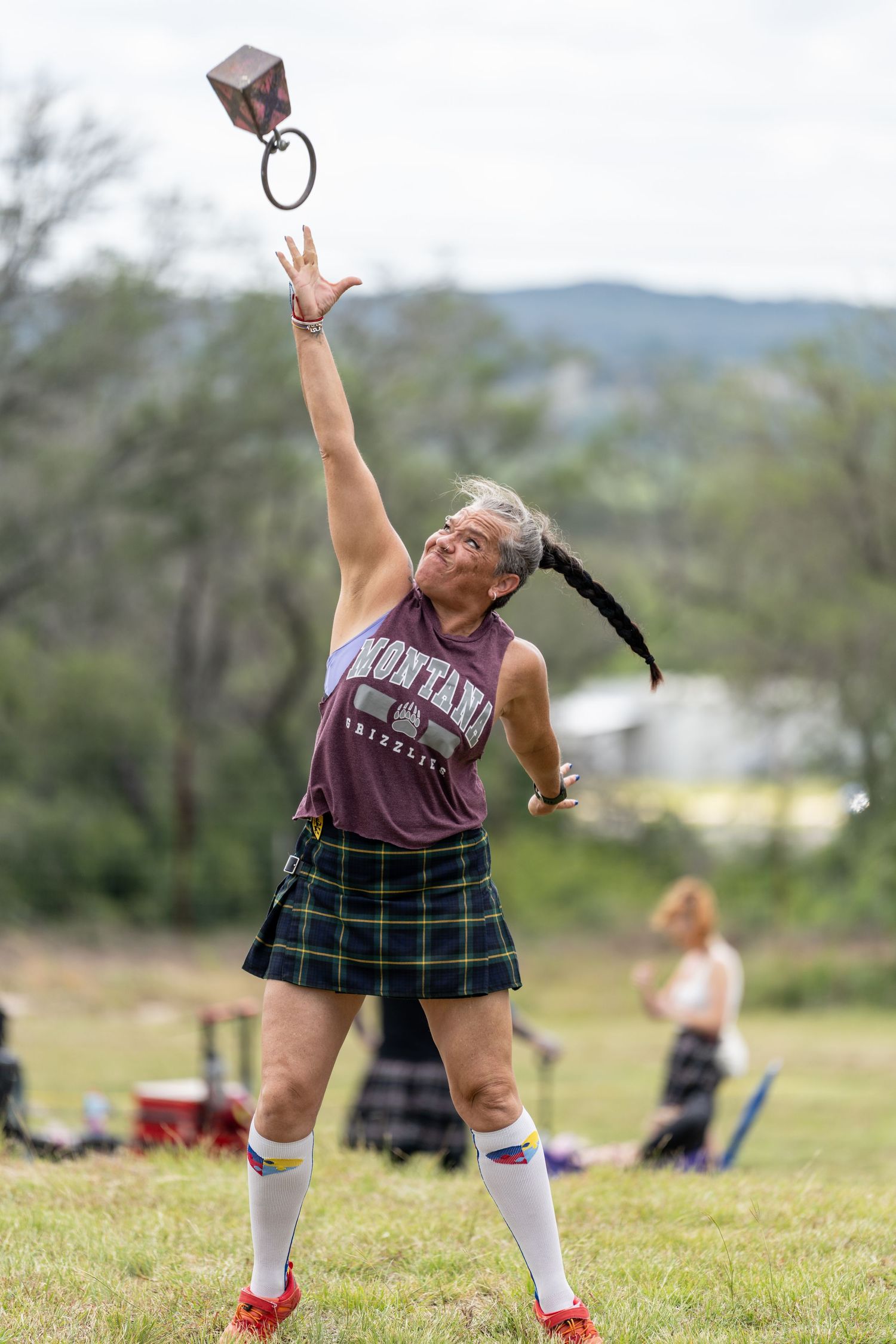 Woman in tartan kilt and Montana shirt tosses weight overhead at Highland Games competition outdoors.