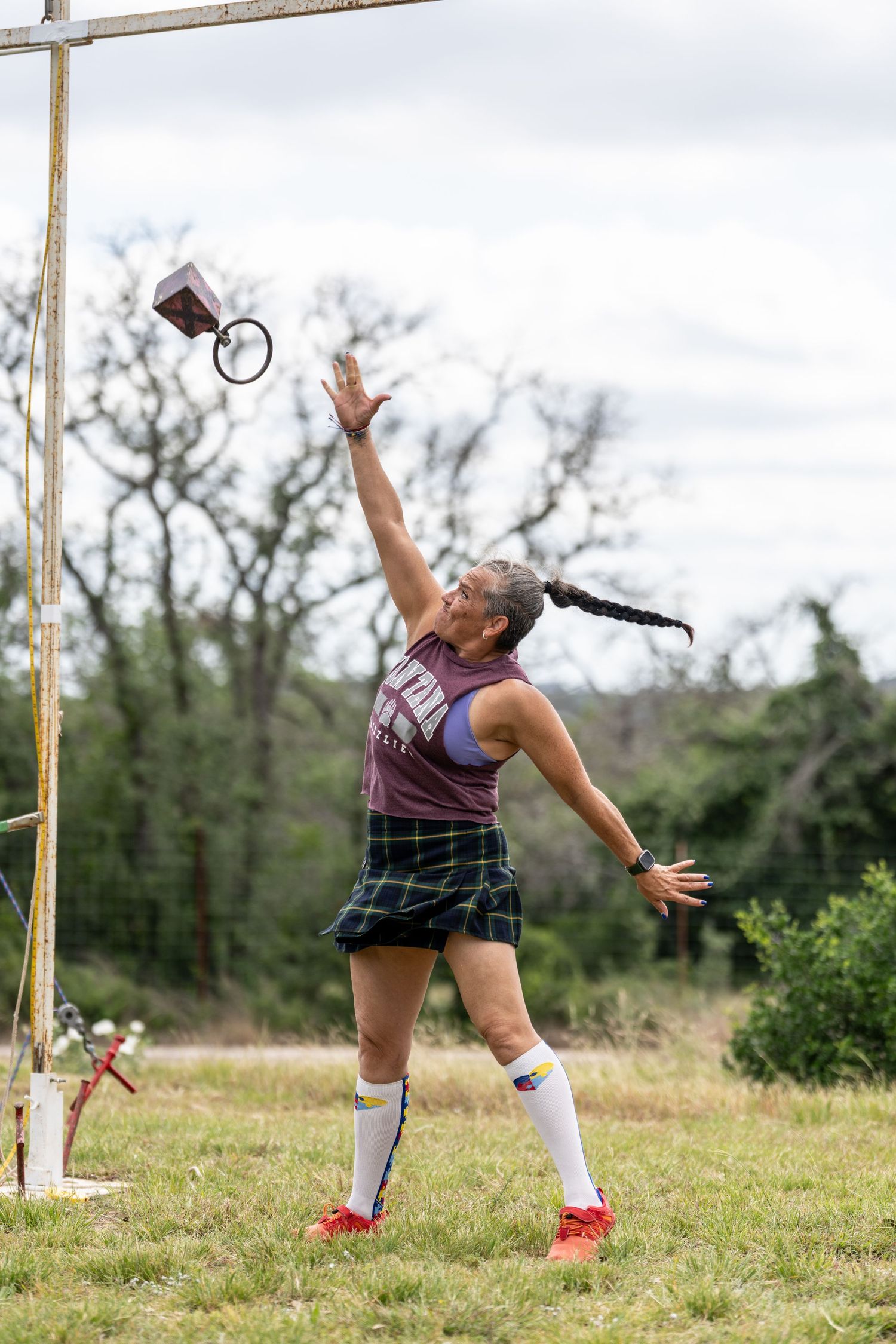 Woman in plaid skirt and white socks throws object skyward during Highland Games competition outdoors.