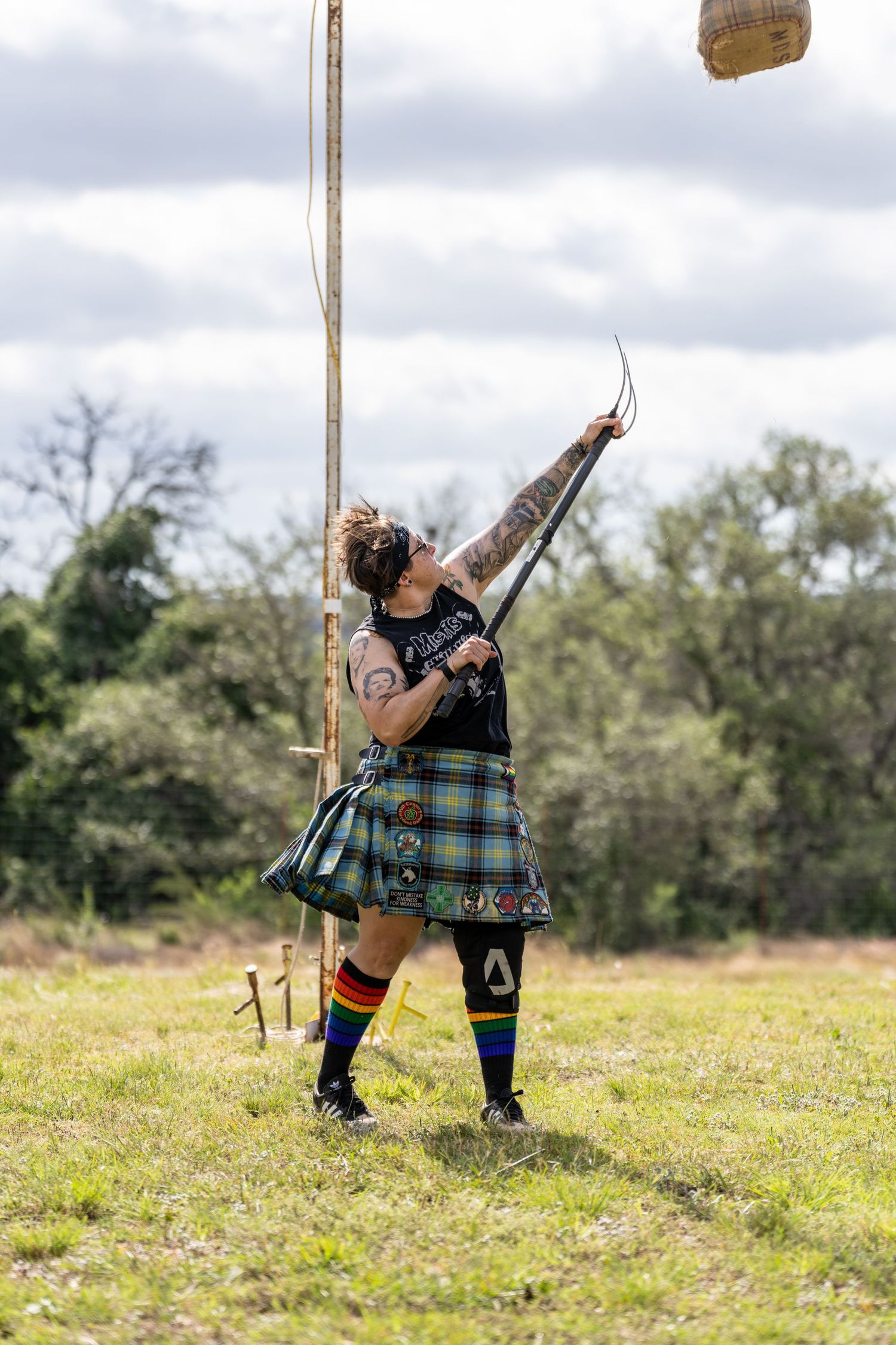 Tattooed archer in tartan kilt shoots arrow skyward at outdoor Highland games competition.
