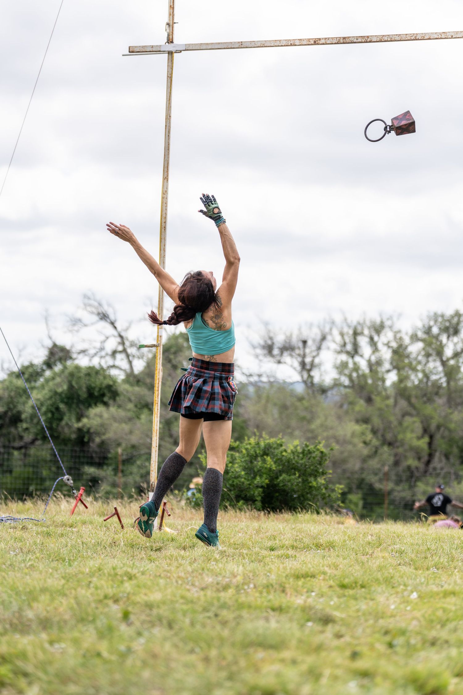 Woman in plaid skirt and knee socks tosses a weight in a Highland Games competition outdoors.
