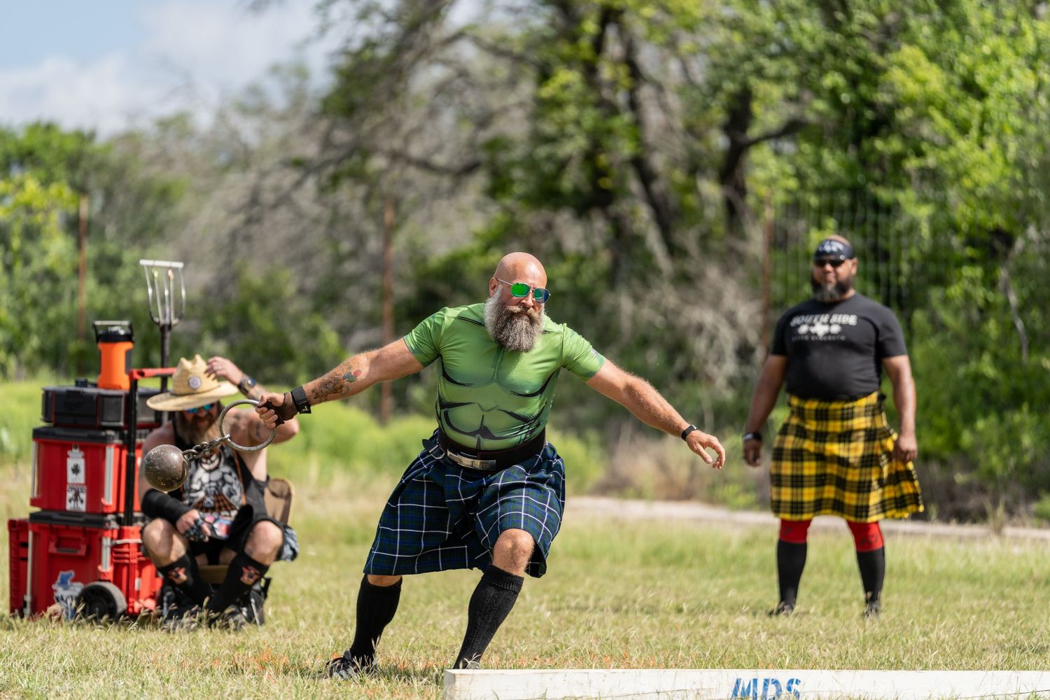 Men in kilts compete in Highland Games athletic events outdoors, with one athlete in mid-throw stance.
