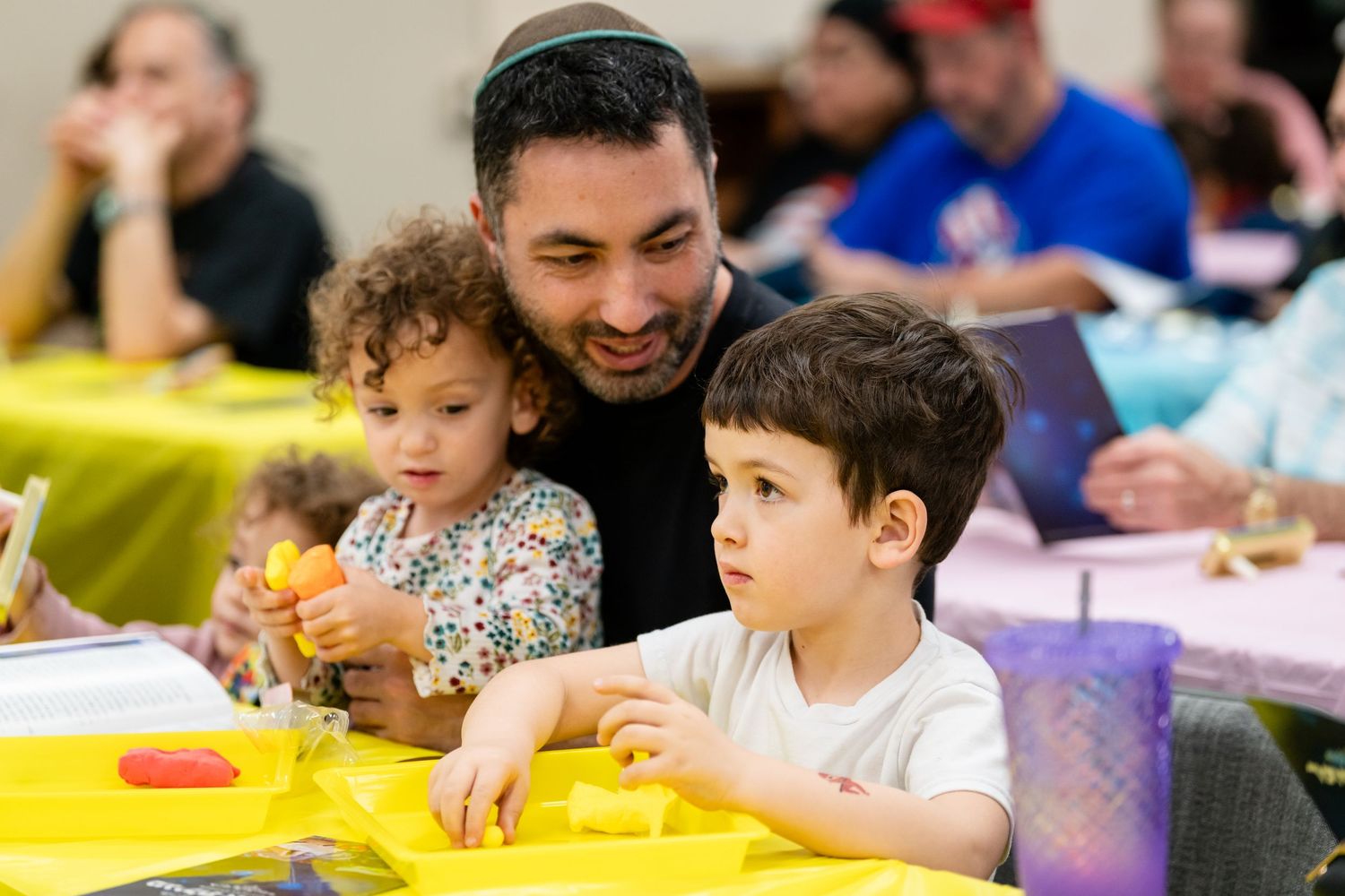 A father smiles with two young children at a yellow table during a festive family craft activity event.