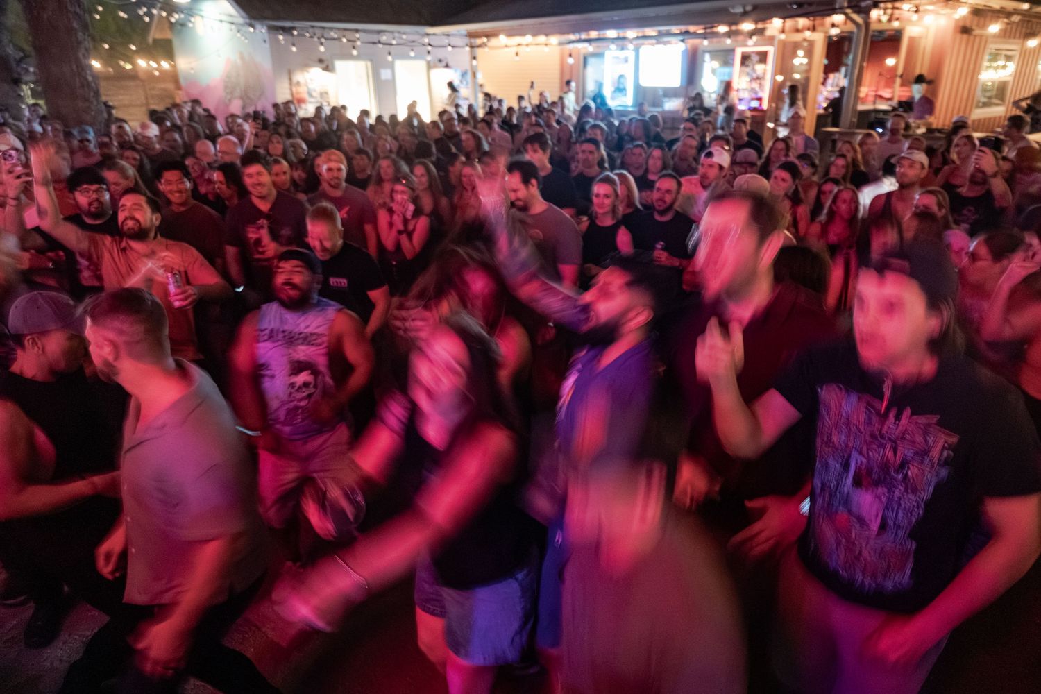 A large energetic crowd dancing at an outdoor concert venue with colorful purple and red lighting.