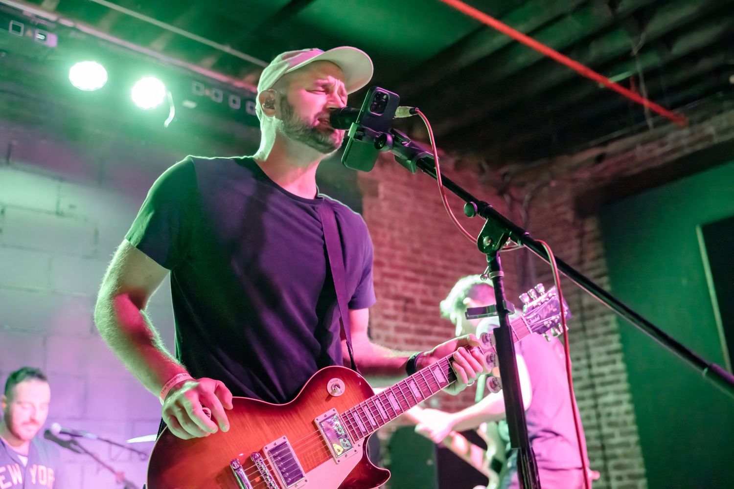 Musician plays red guitar while singing into microphone on stage with brick wall backdrop.