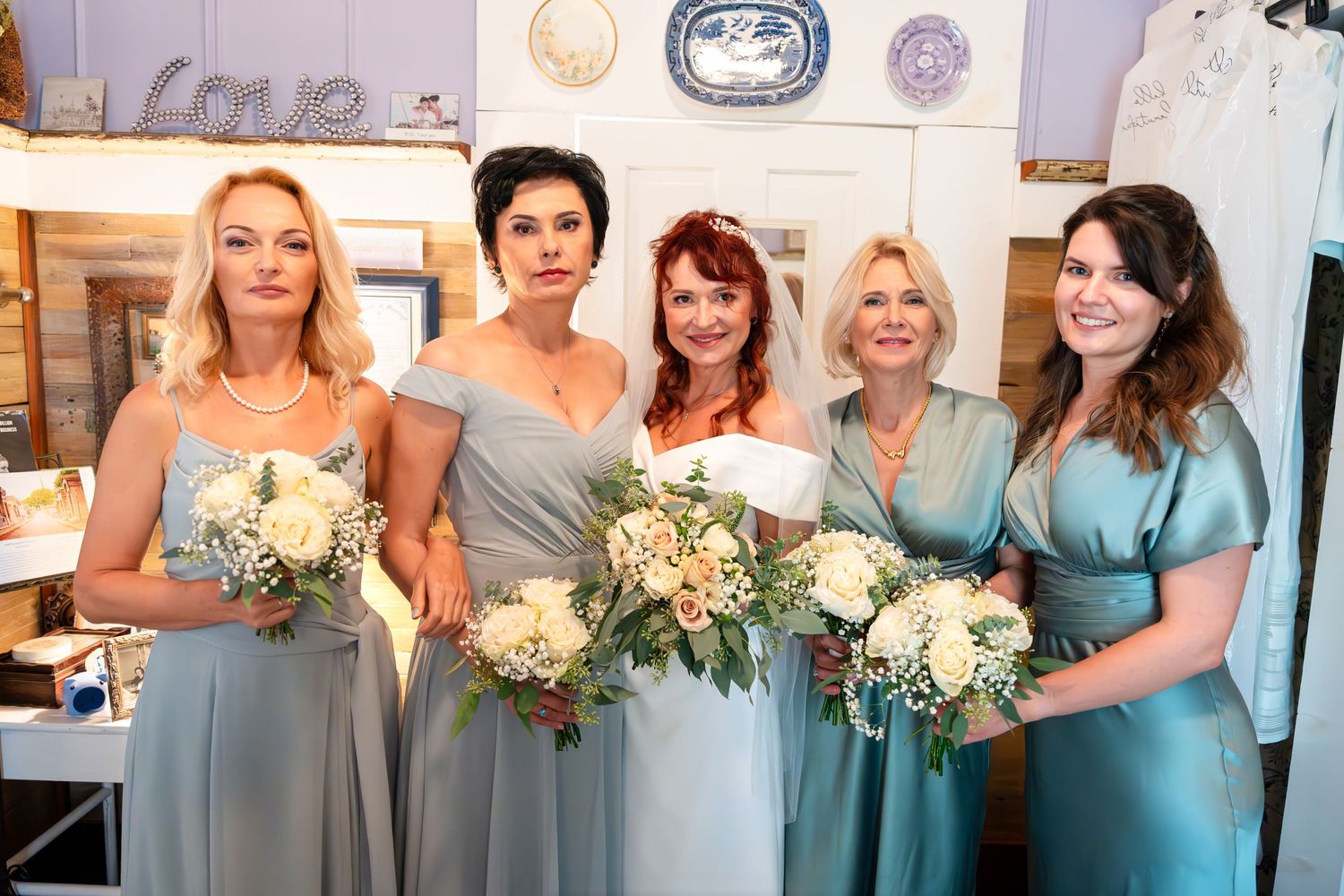 Bridal party poses in coordinating blue and teal dresses holding elegant white floral bouquets.