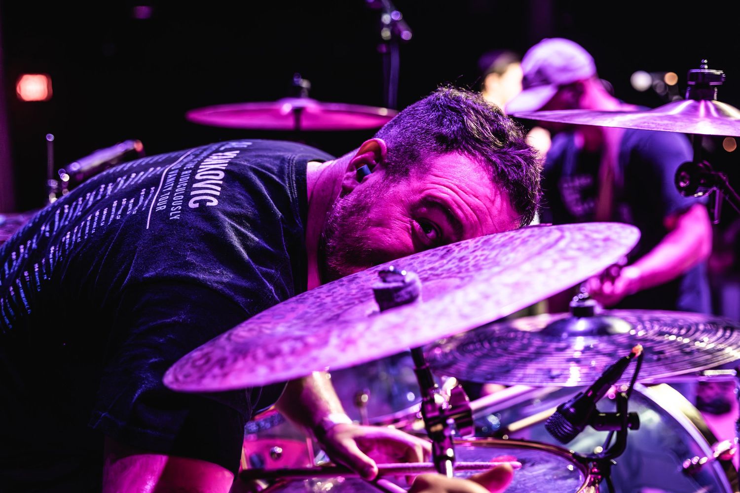 Dramatic shot of drummer bent over cymbals during intense performance bathed in purple light.