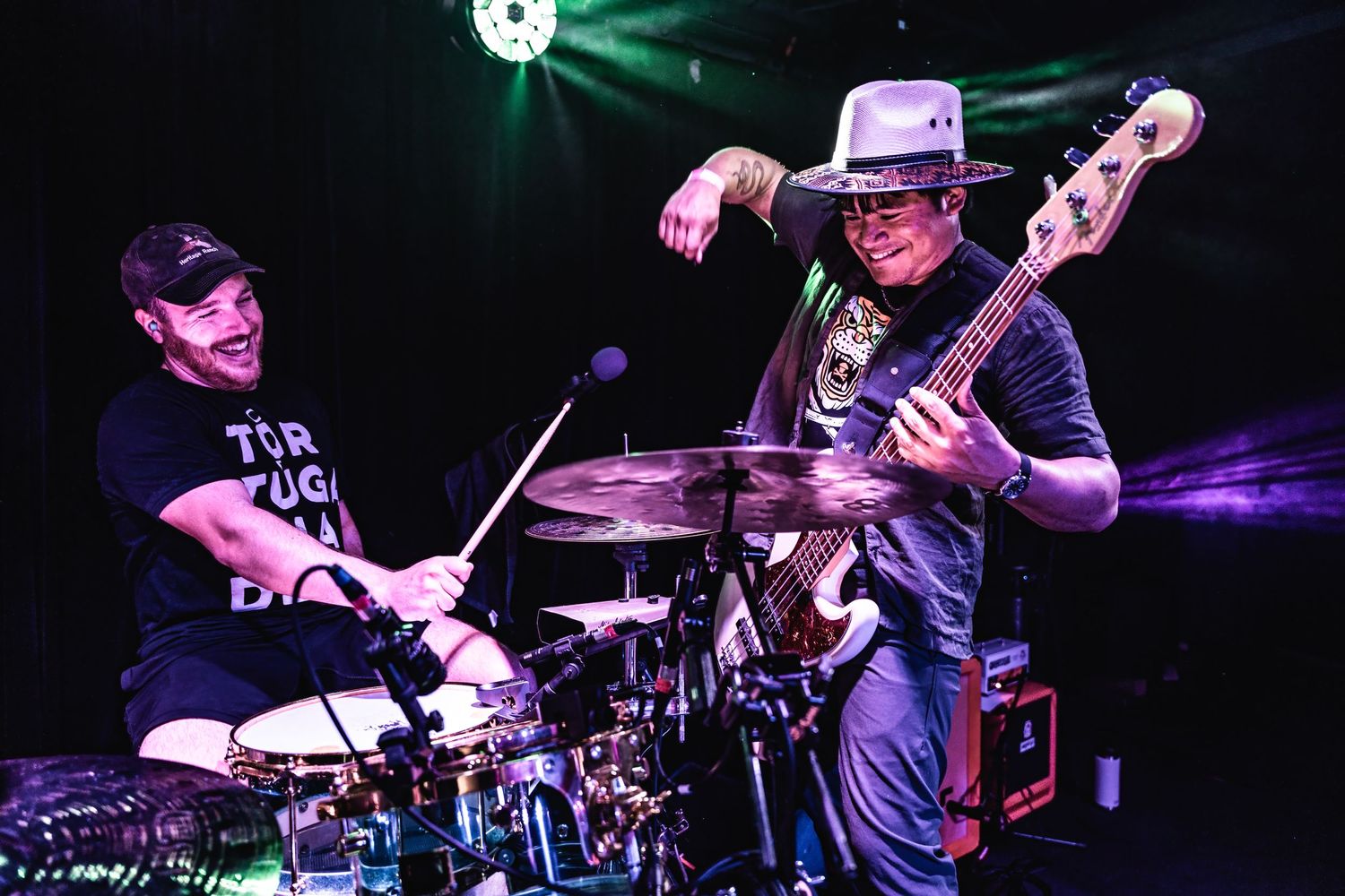 Drummer and bassist perform together under dramatic stage lighting during a live show.