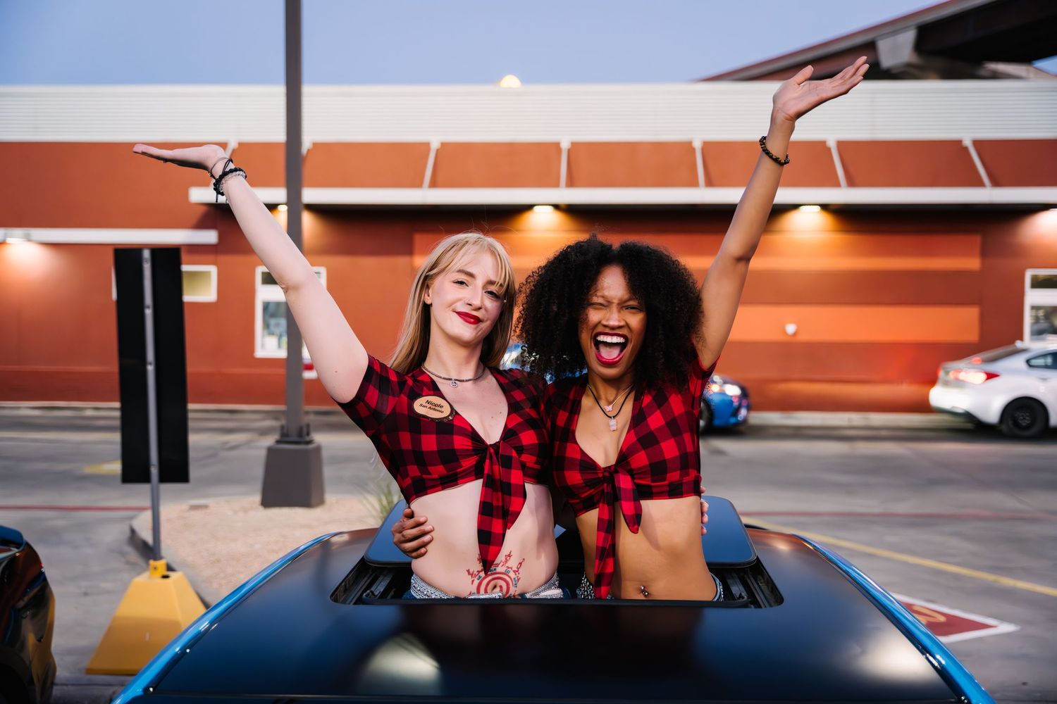 Friends in red plaid tops pose energetically through sunroof of car at outdoor event.