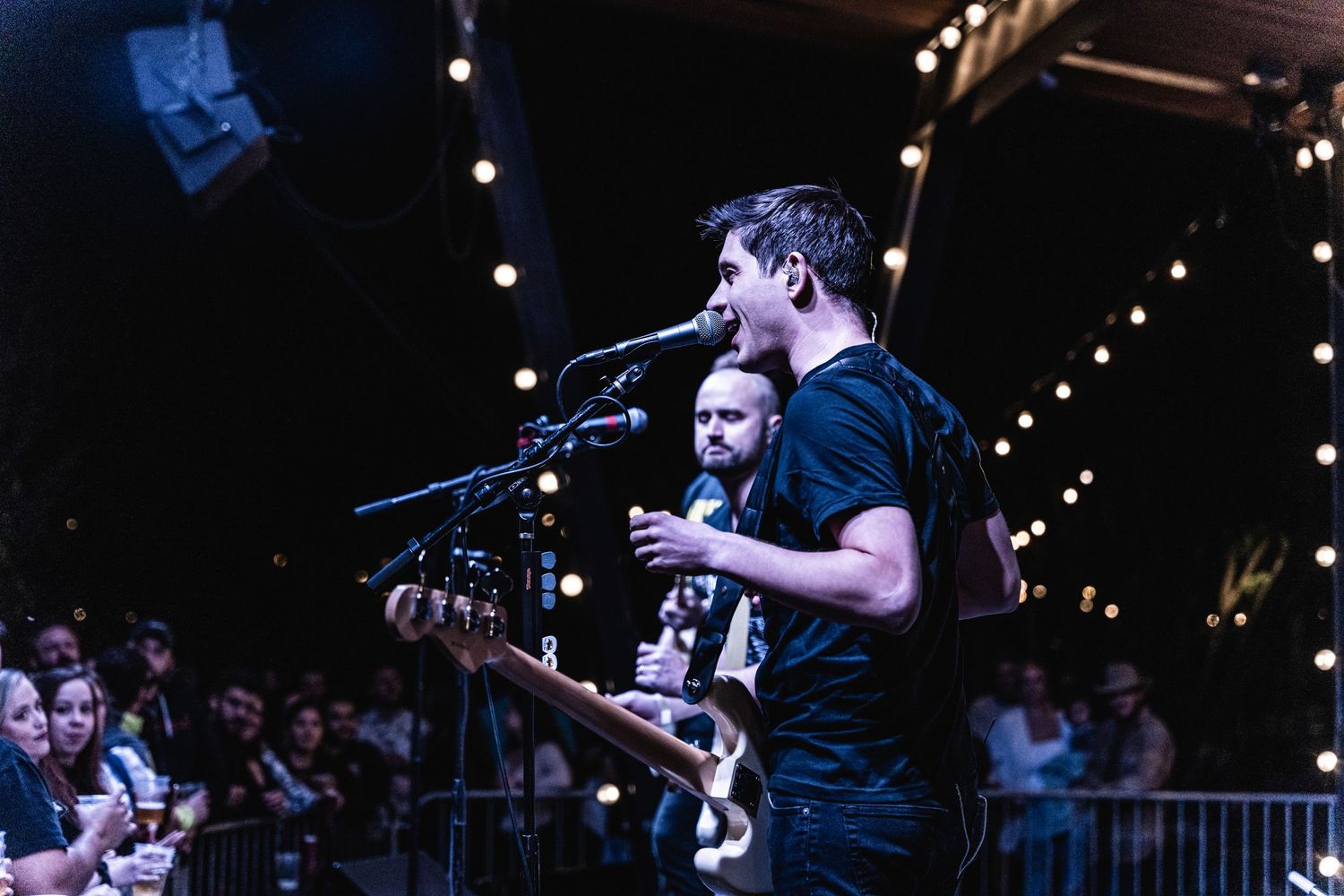 Musicians perform on stage with string lights and crowd barriers visible.