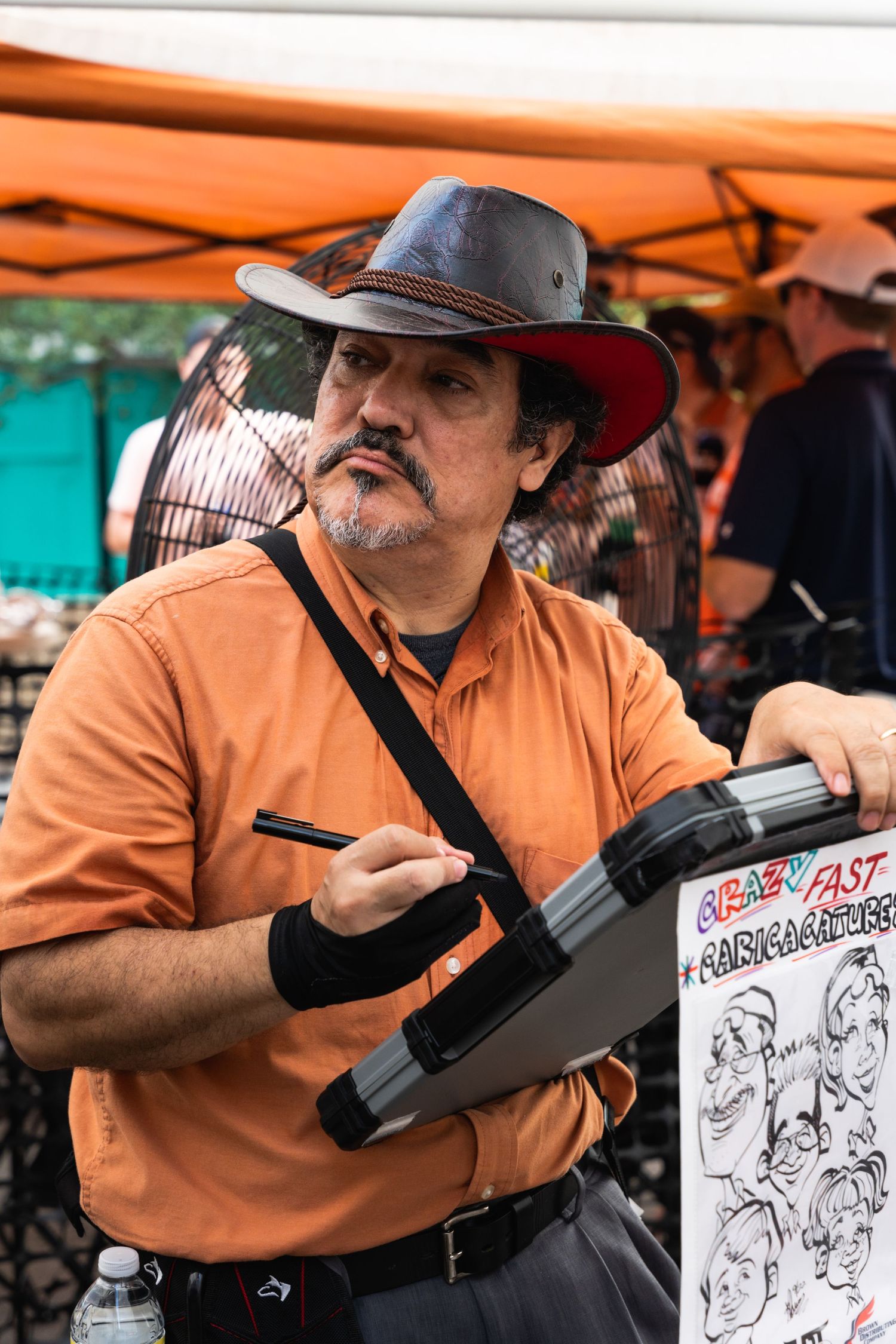 Artist wearing cowboy hat and orange shirt sketching caricatures at outdoor festival.