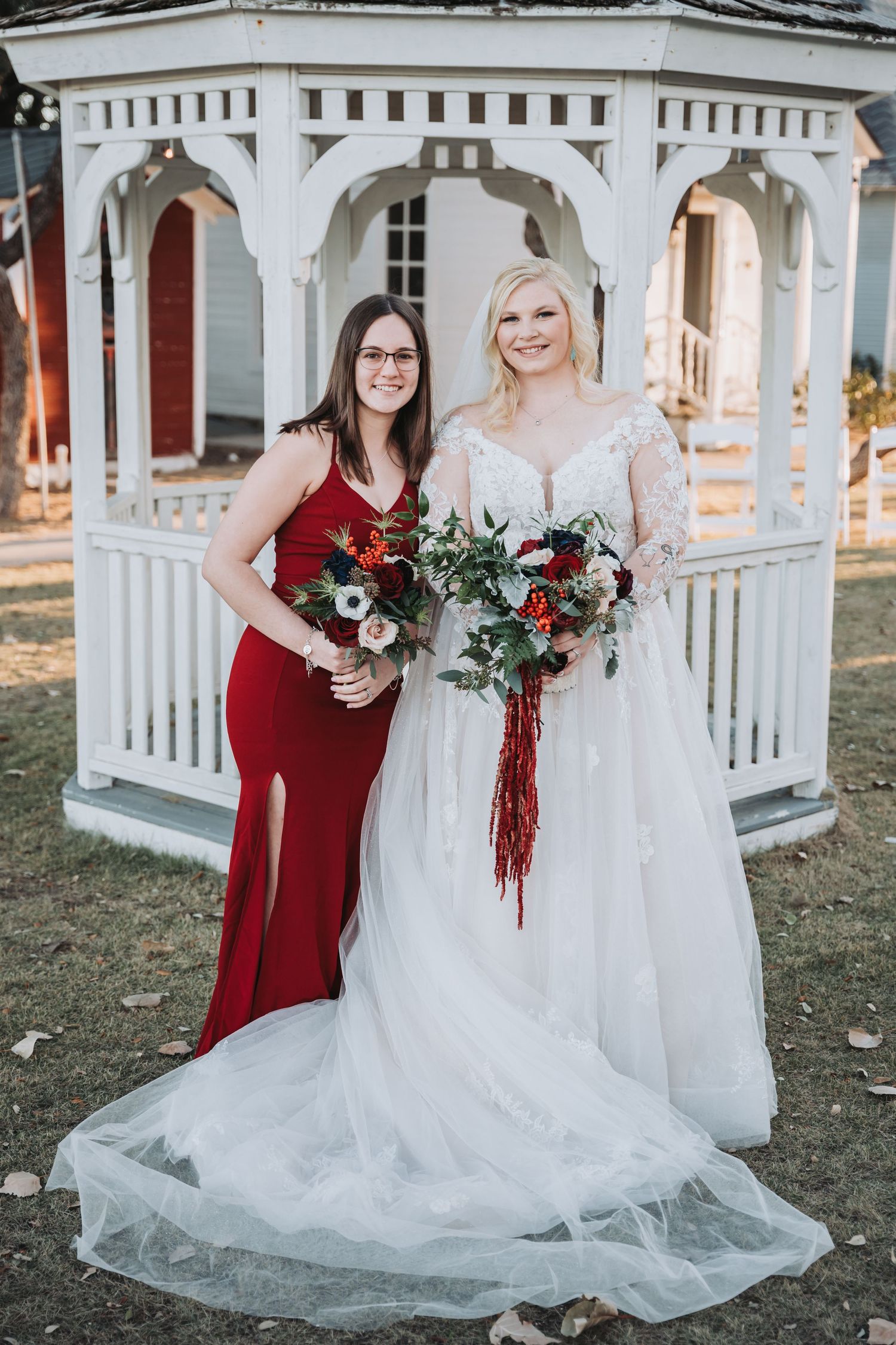 Two attendants hold floral bouquets while posing in front of a white gazebo.