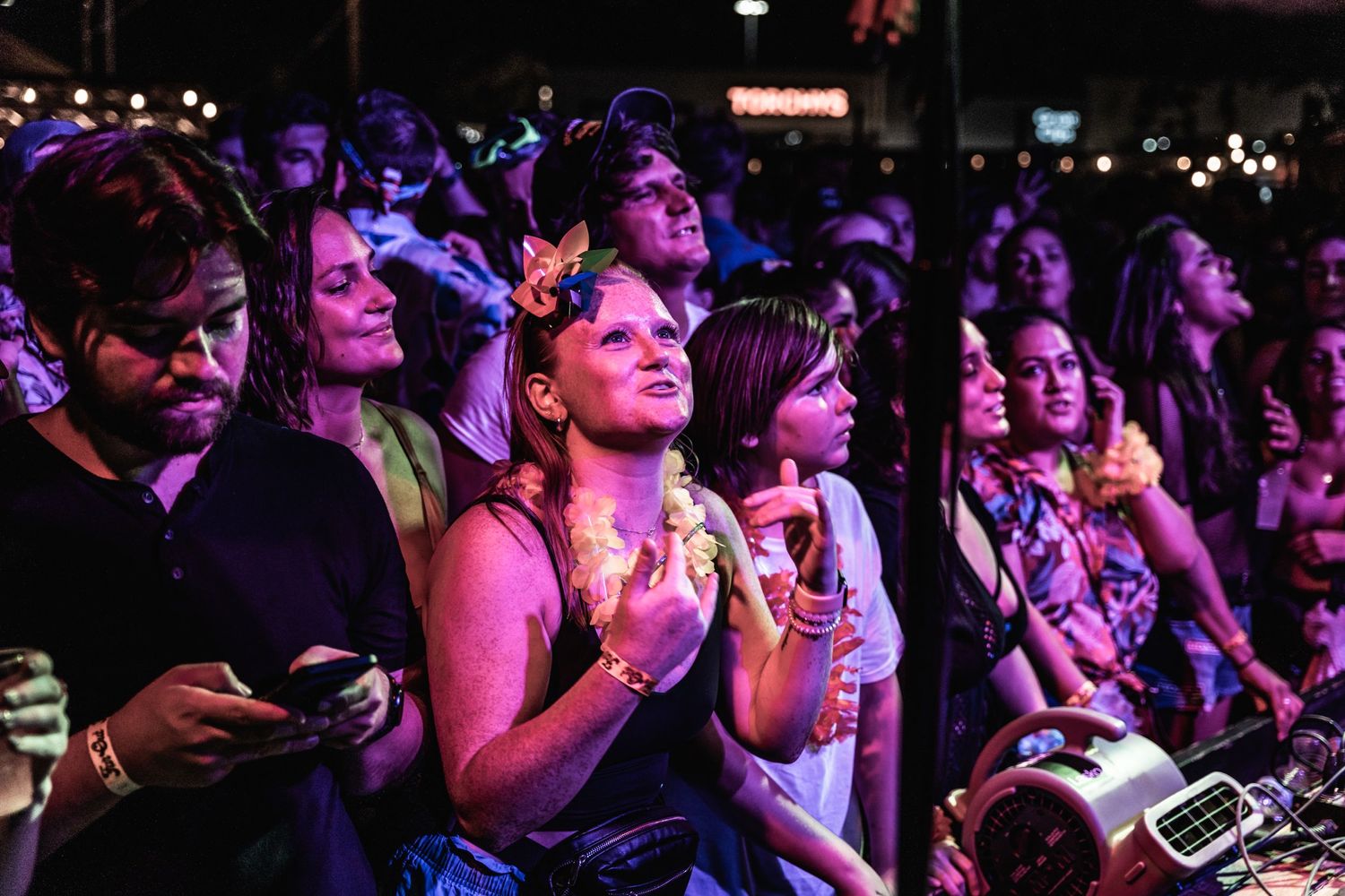 Concert crowd watches performance with phones and drinks in hand under purple lighting.