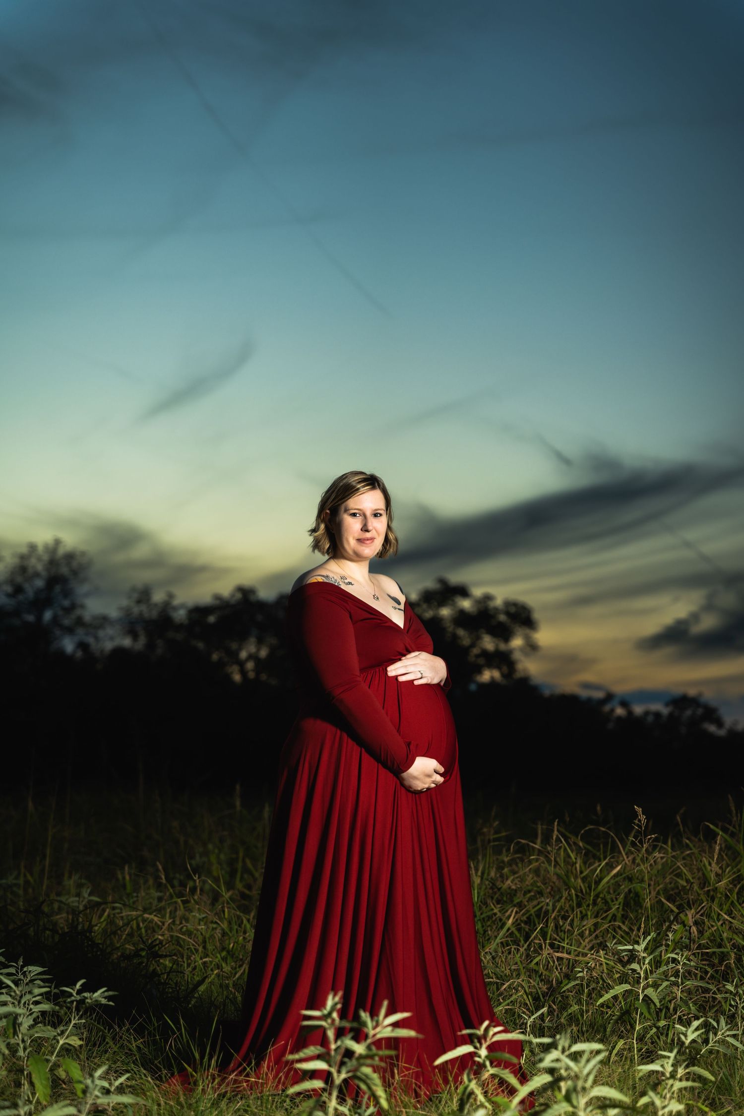 Dramatic sunset maternity portrait with woman in red dress standing in tall grass.