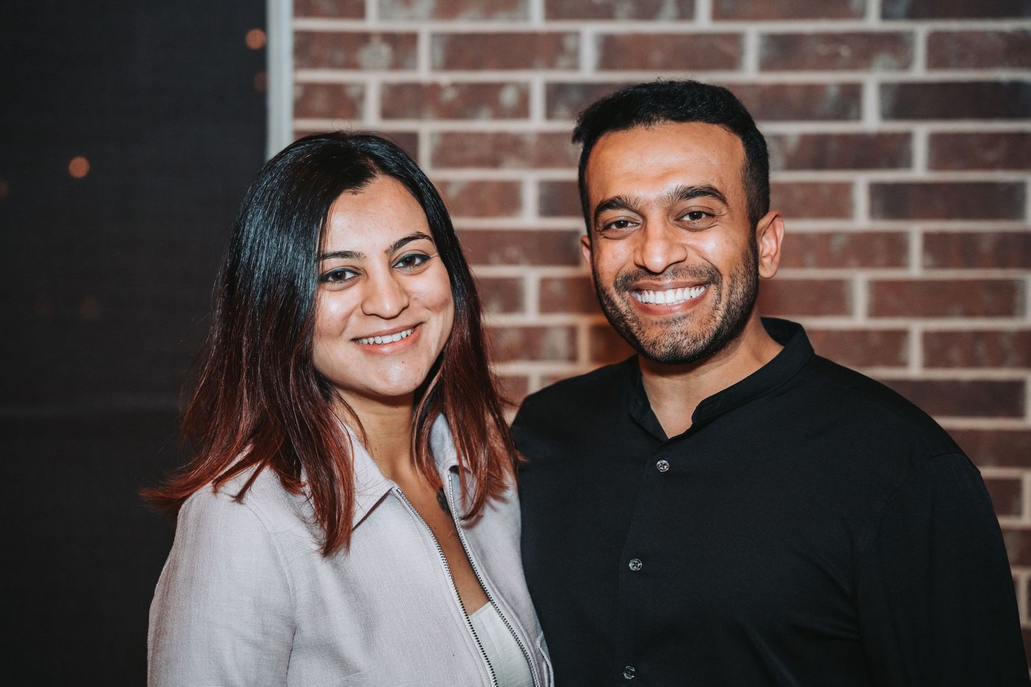 Couple smiles warmly against brick wall background at evening celebration.