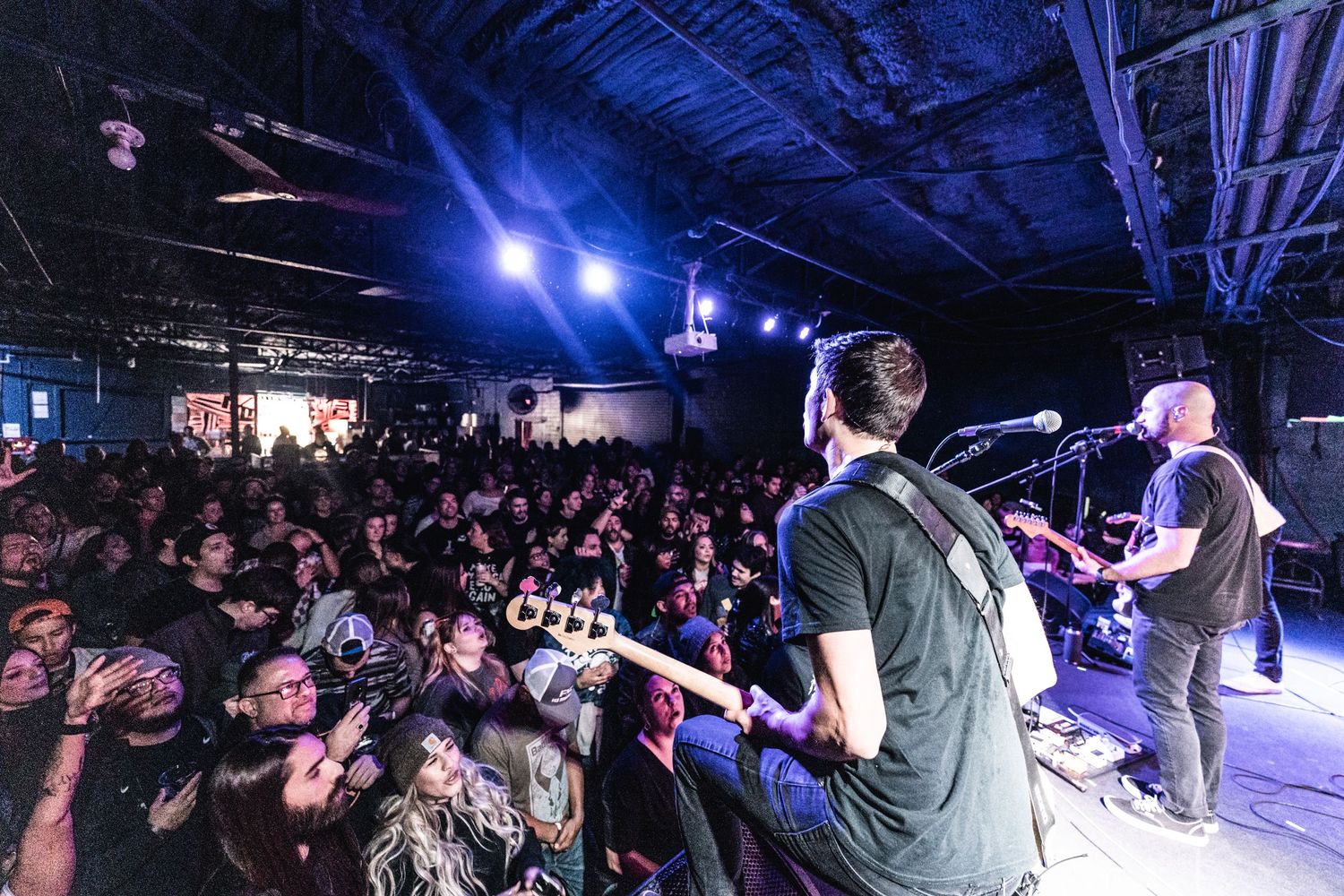 Wide angle view of band performing on stage with packed audience beneath blue stage lights.
