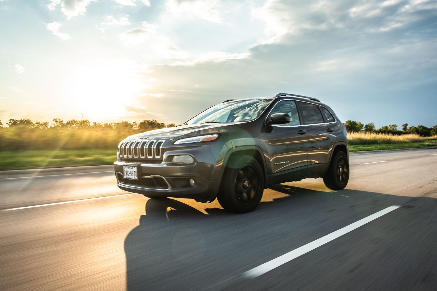 Gray Jeep Cherokee with custom wheels driving at sunset on empty highway.