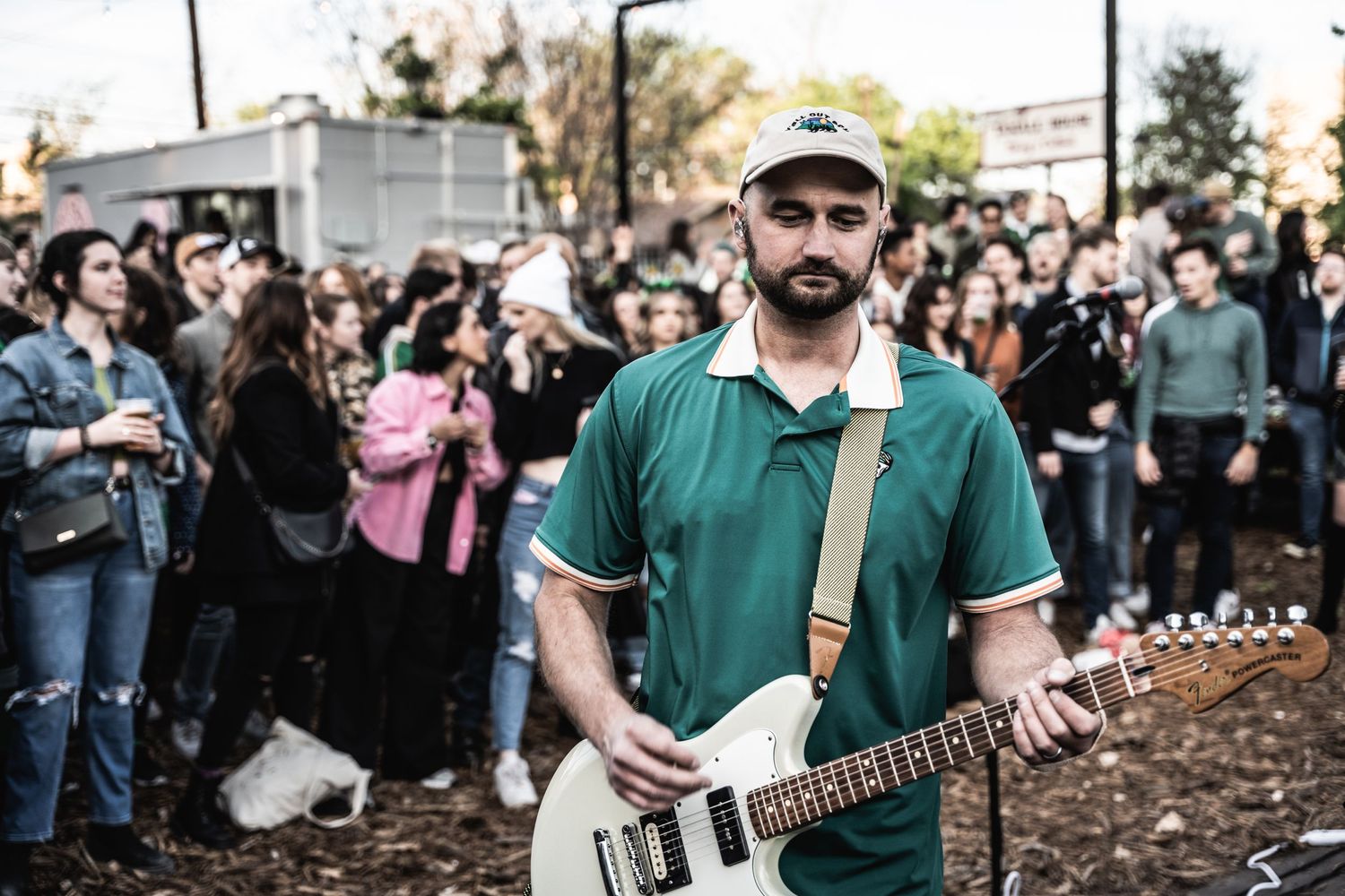 Guitarist in green polo performs at outdoor event with large crowd gathering behind.
