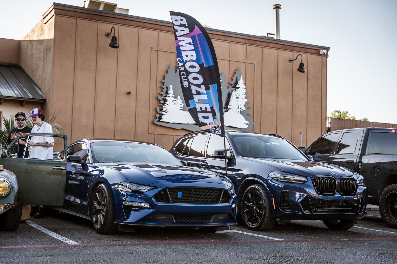 Two performance cars, a blue Mustang and black BMW, displayed at a car meet with a banner flag.