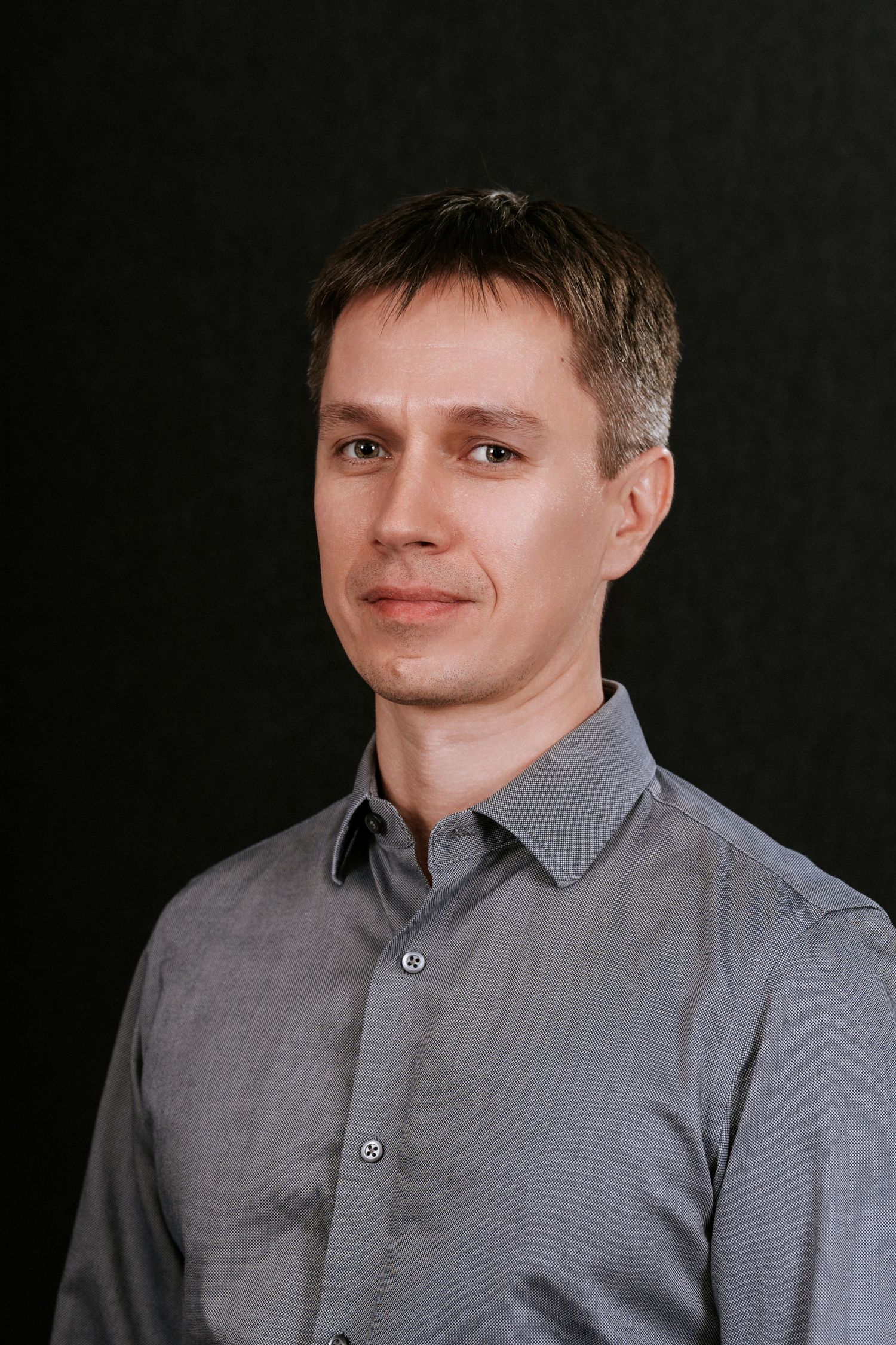 Professional headshot of a person wearing a grey button-up shirt against a dark background.